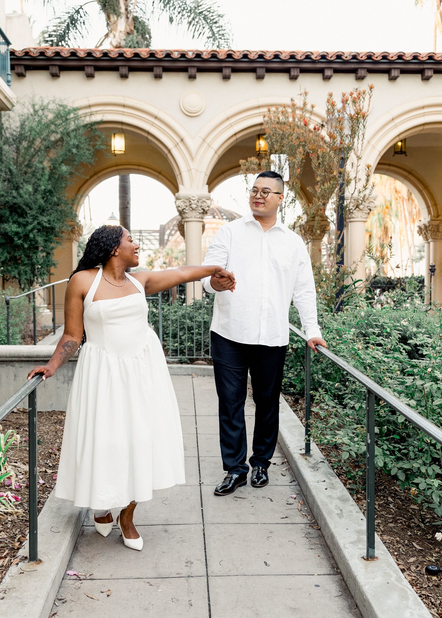 Couple holding hands and walking down a path in front of Spanish-style arches in San Diego
