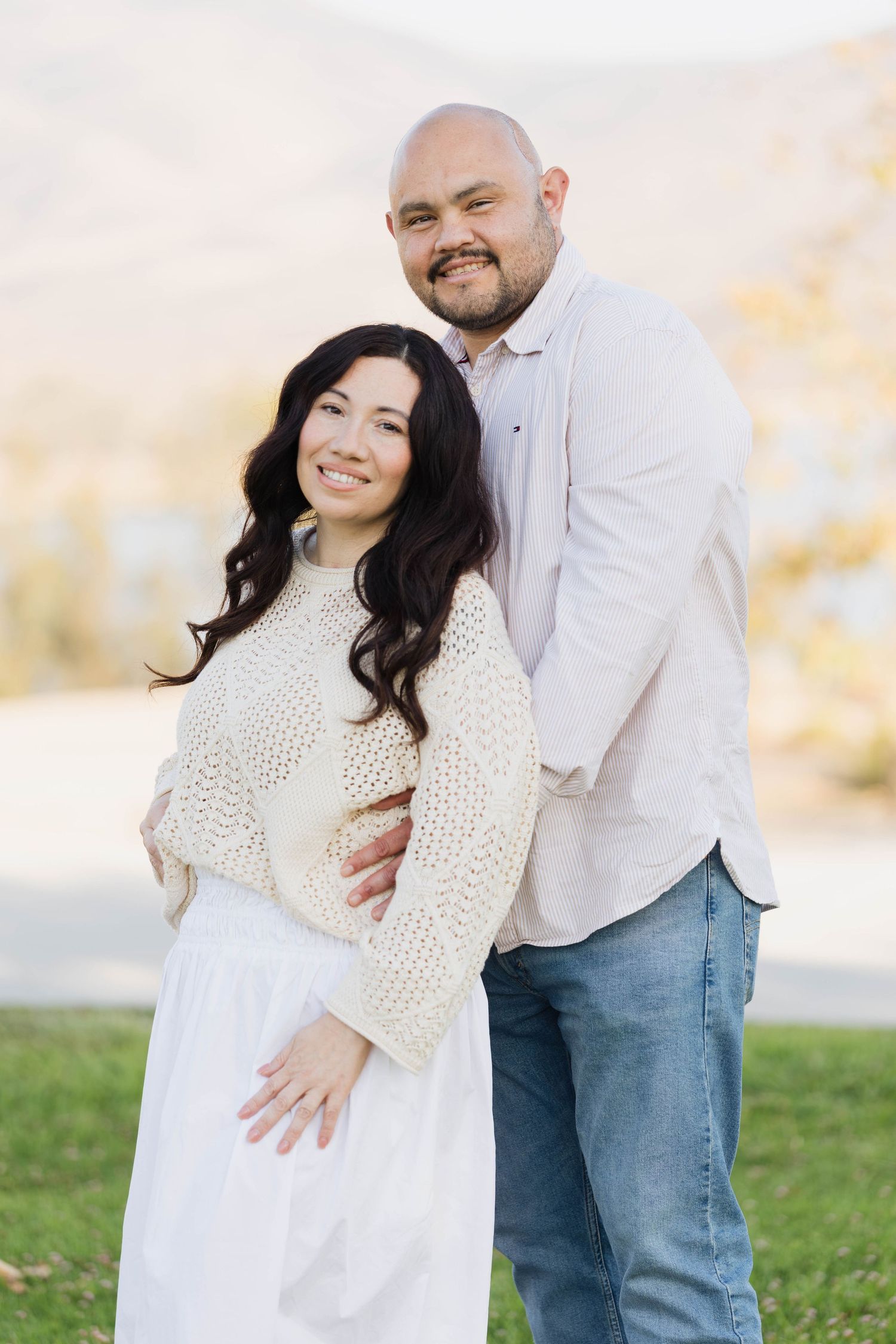Husband embracing his smiling wife from behind on a grassy hillside during an outdoor couples session in San Diego