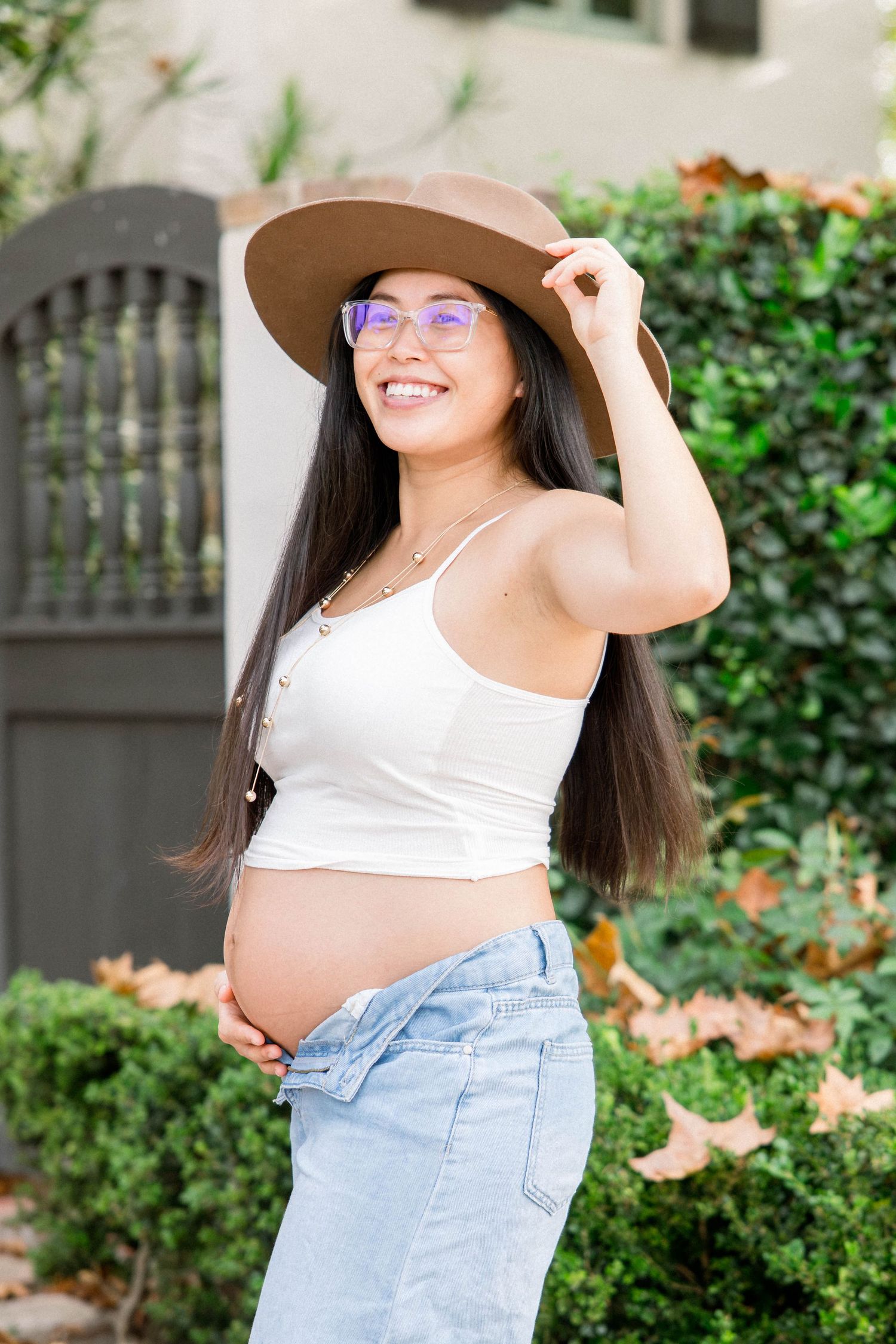 Smiling pregnant woman in a tan hat and glasses posing with a hand on her baby bump during a San Diego maternity session