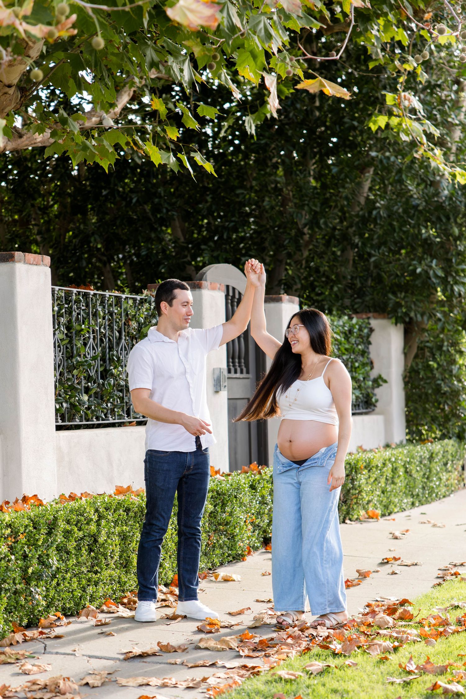 Husband twirling his pregnant wife on a leaf-covered sidewalk during an outdoor maternity photoshoot in San Diego