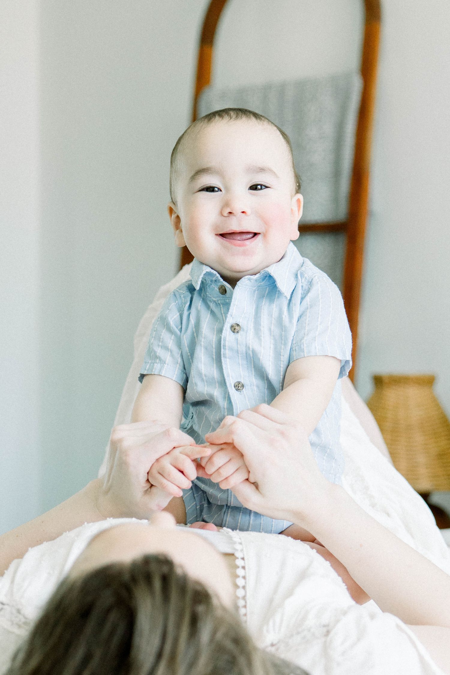 Smiling baby in a blue button-up shirt held up by his mother during a light, airy motherhood session