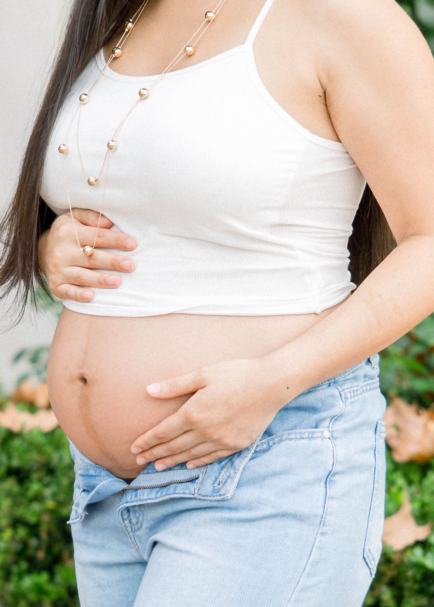 Pregnant woman in a white top and jeans gently holding her baby bump during an outdoor maternity session