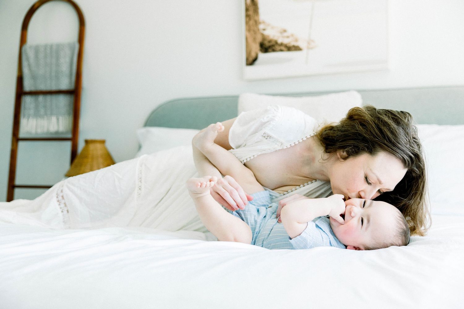 Mother in a white dress kissing her baby boy on a bed during a bright, airy in-home motherhood session in San Diego