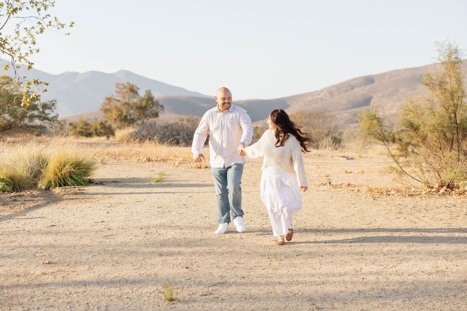 Couple walking hand in hand down a dirt path with mountains behind them during a sunset couples session in San Diego