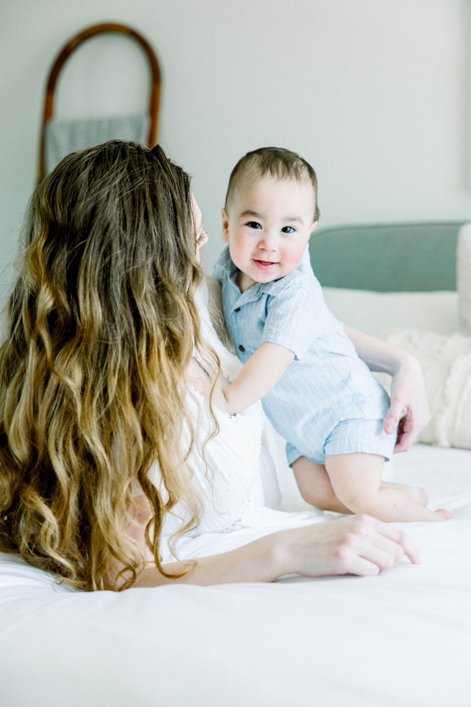 Baby sitting up on a bed and smiling at the camera while his mother looks on during a bright in-home motherhood session in San Diego