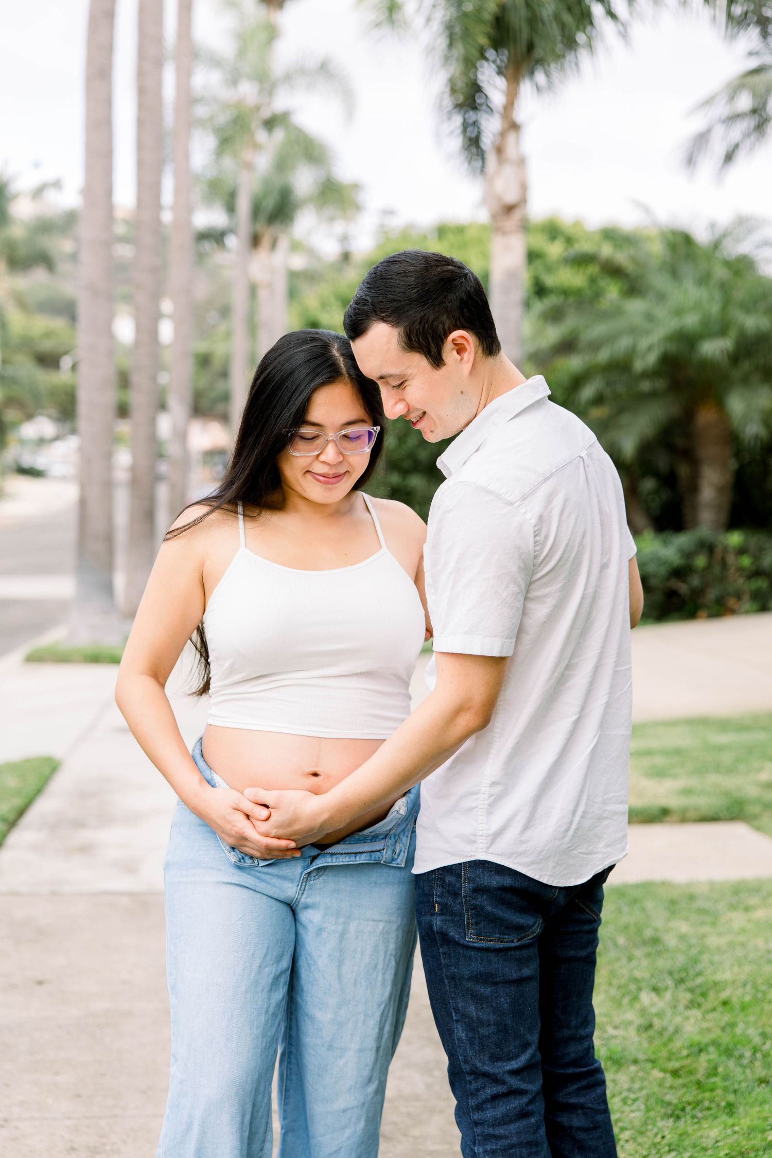 Expecting couple touching foreheads and cradling the baby bump under palm trees during a San Diego maternity session