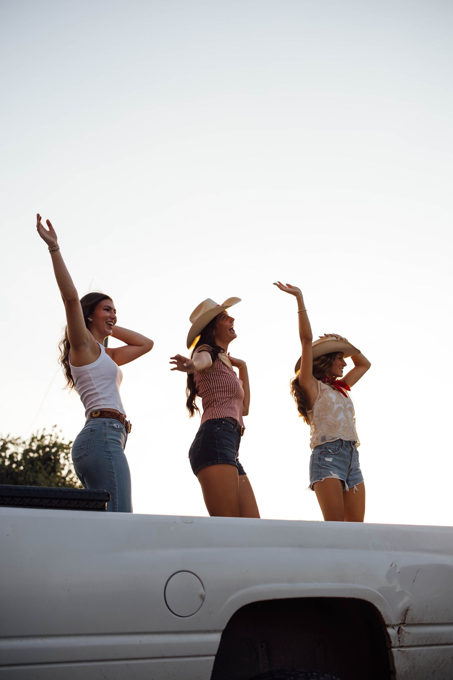 Three women in cowboy hats dance joyfully standing in a pickup truck bed at sunset.