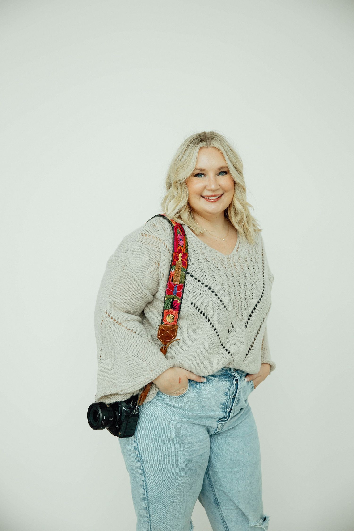 Smiling blonde female photographer in beige sweater and jeans holding camera with colorful strap.