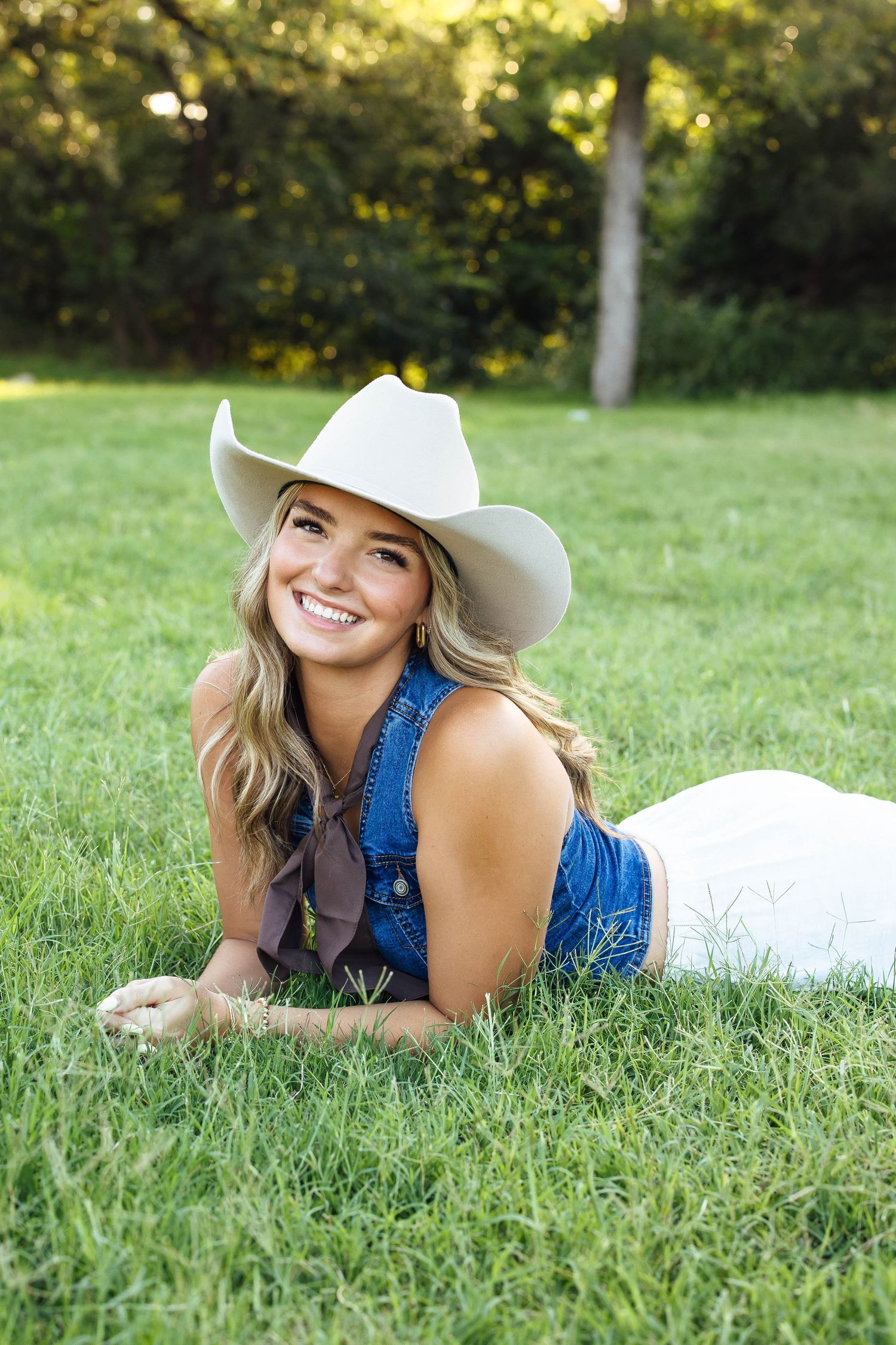 Smiling blonde girl in denim overalls and white cowboy hat lying on green grass.