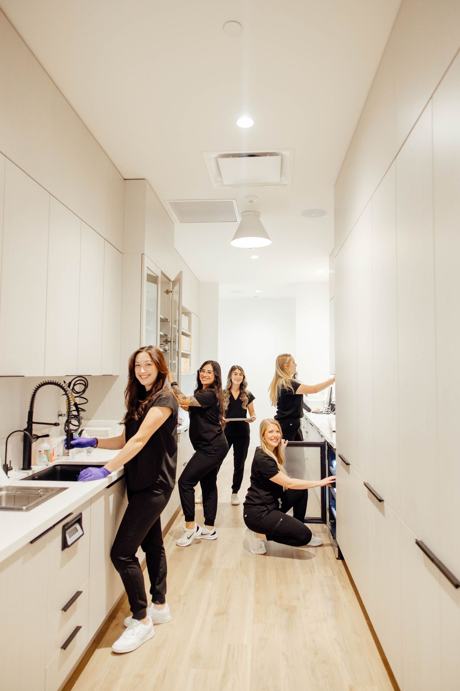 Dental team of five women in black scrubs posing playfully in a modern white kitchen hallway.