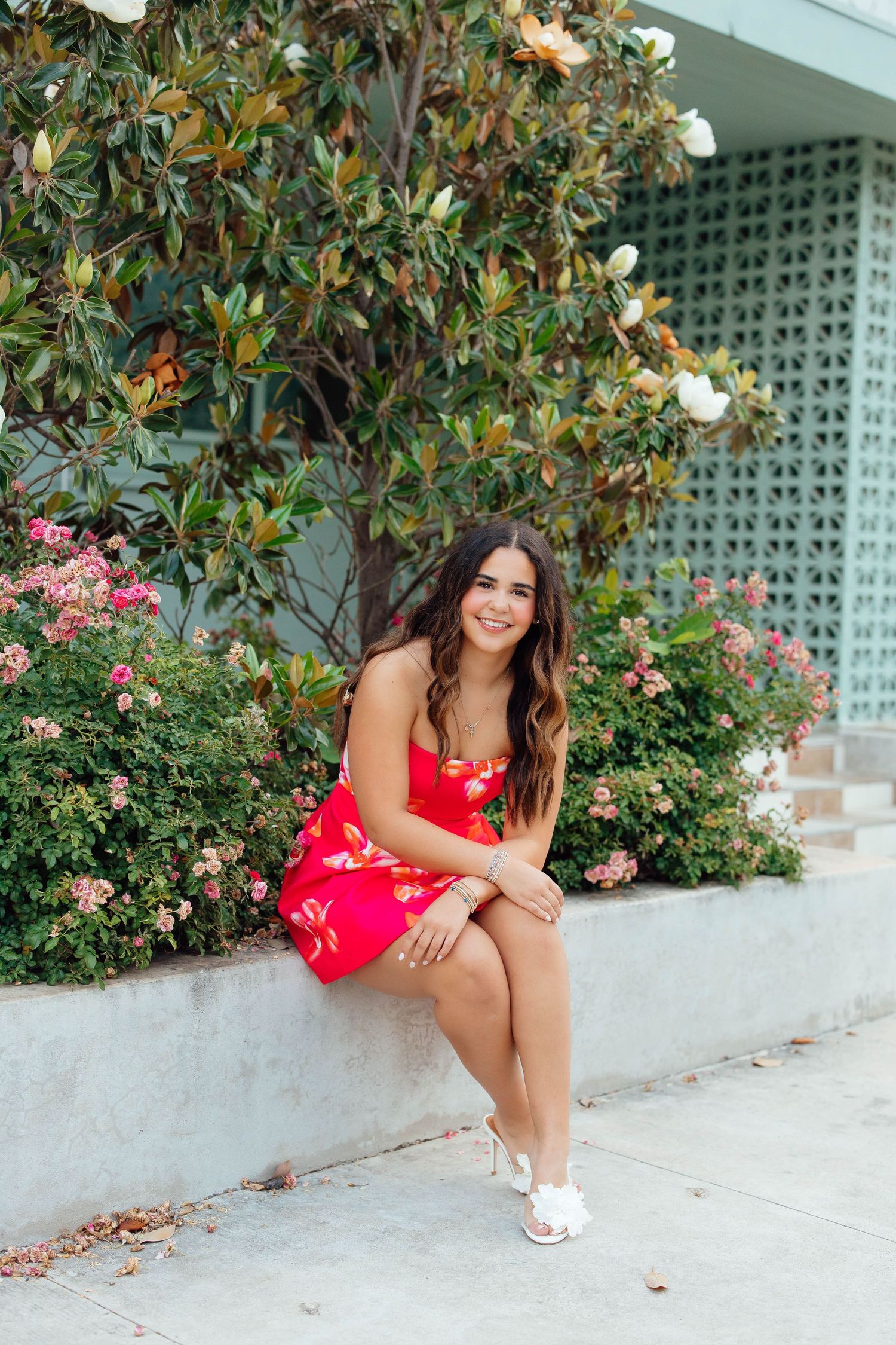Brunette woman in red floral dress sitting on stone ledge surrounded by blooming flowers and greenery.
