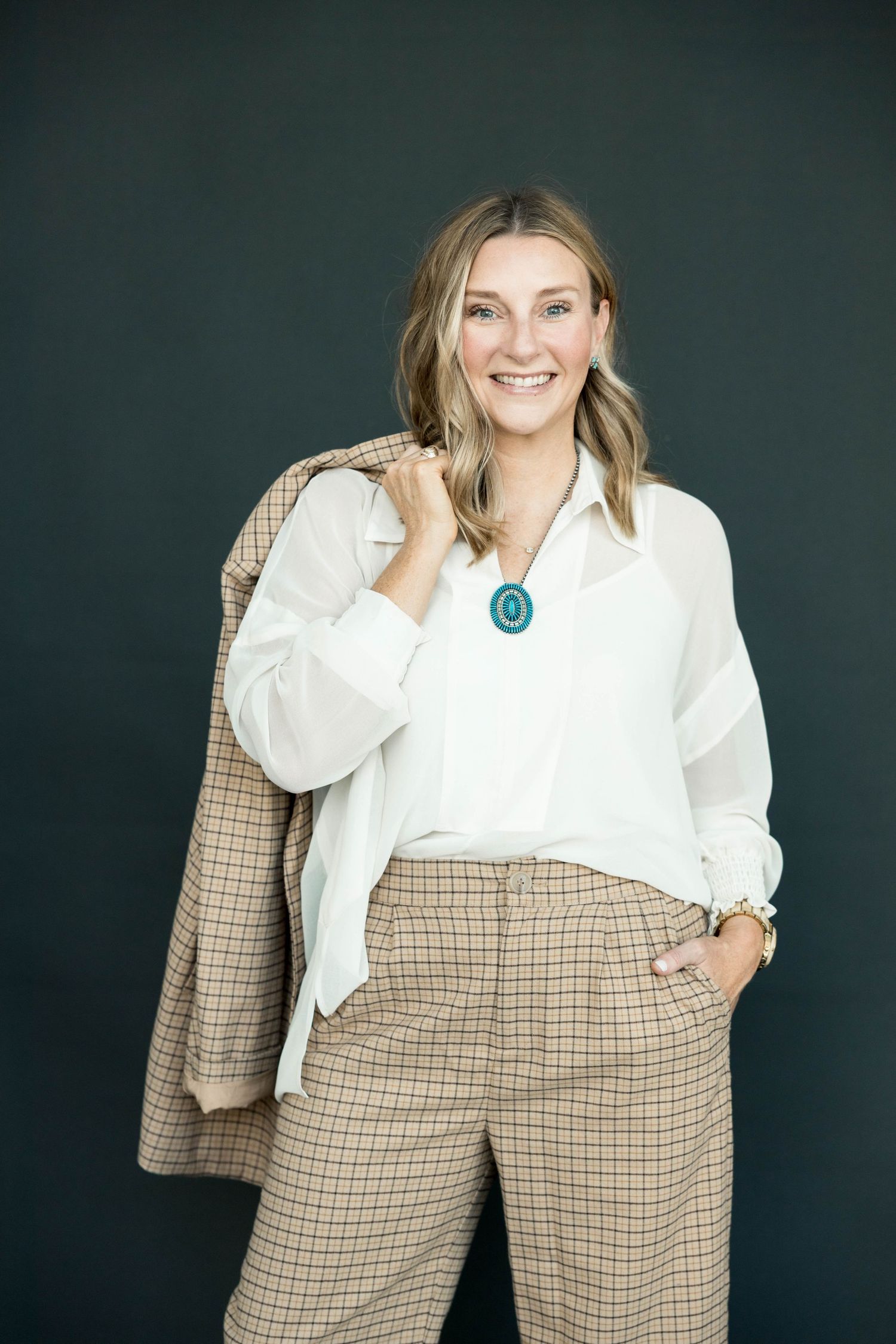 Smiling blonde woman in white blouse and plaid trousers with turquoise necklace against dark green background.