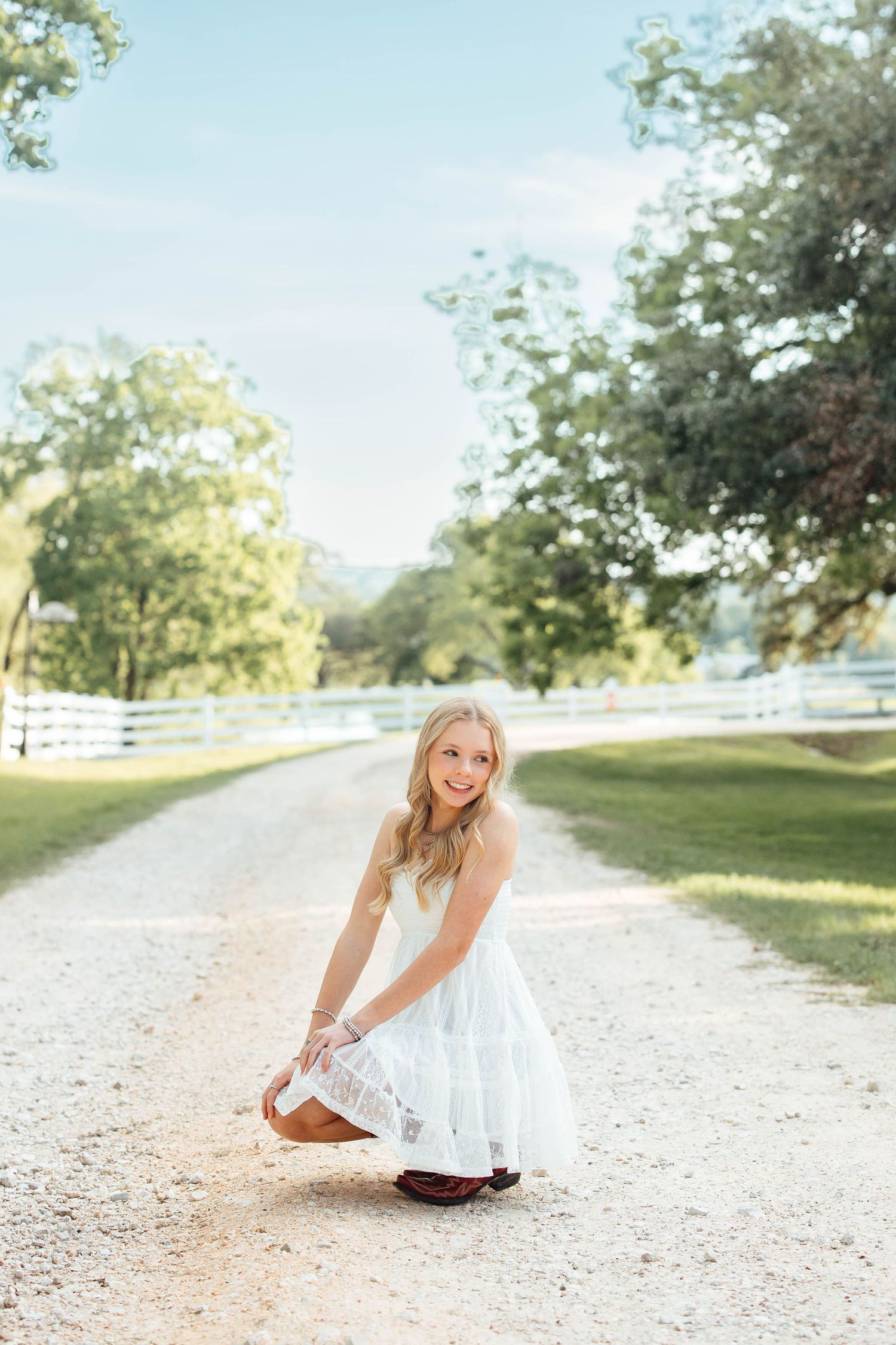 Blonde girl in white dress crouching on sunny gravel path flanked by green trees and white fence.