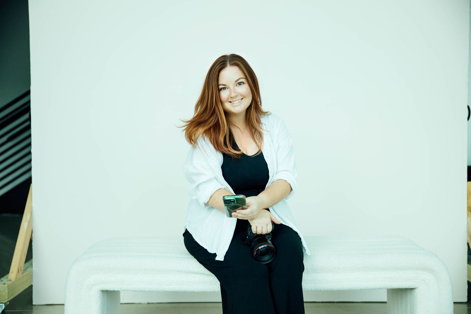 Smiling redhead woman in white blazer sitting on bench holding camera against white background.