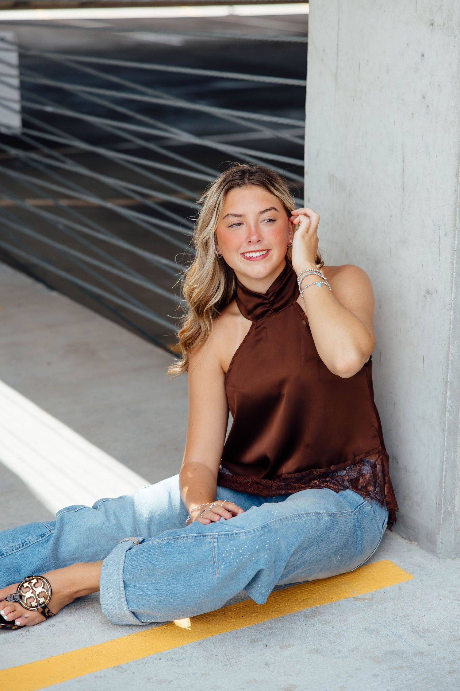 Smiling blonde woman in brown satin halter top and jeans sits relaxed in a modern parking garage.