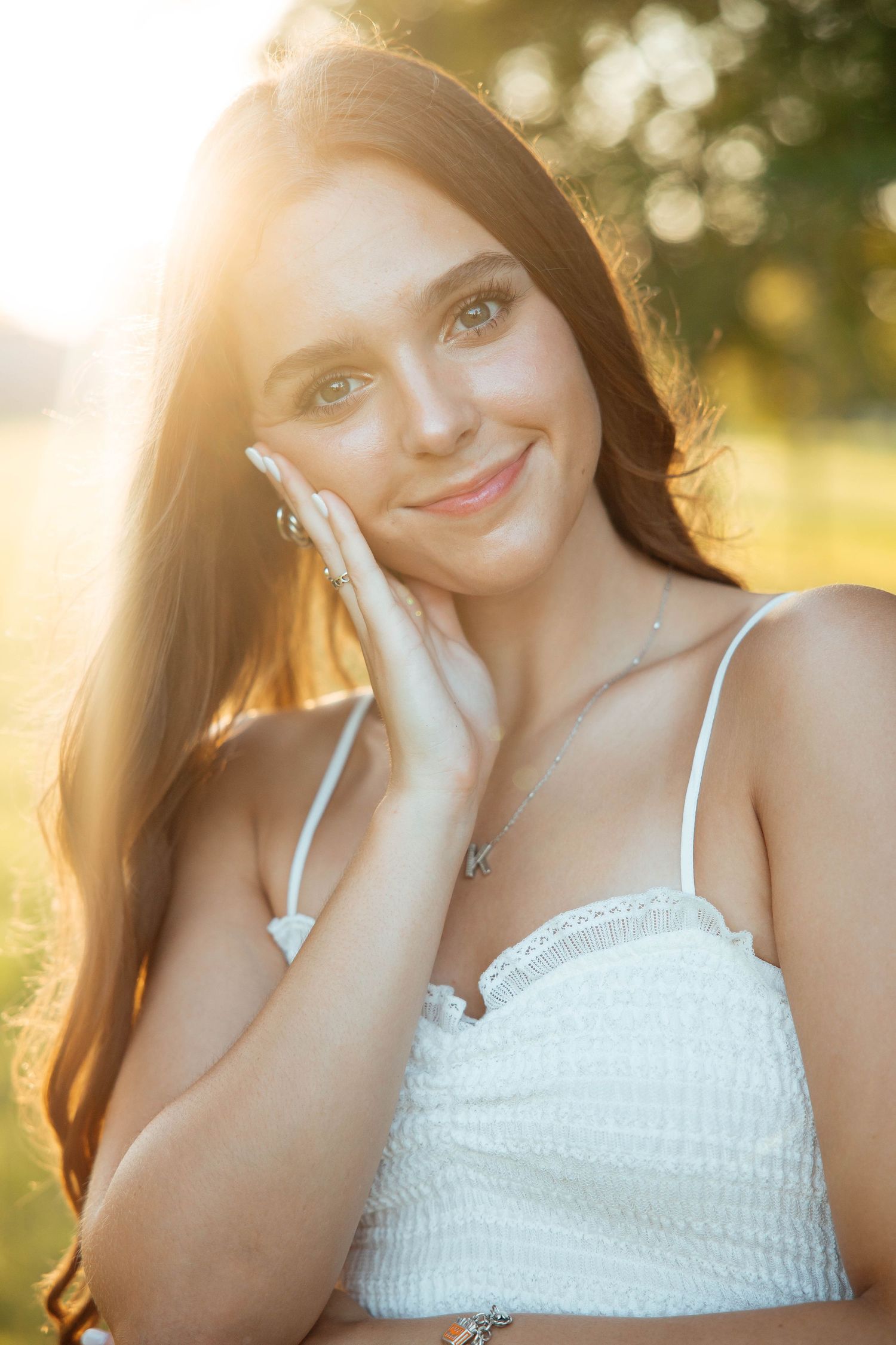 Beautiful brunette teen girl in white dress smiling softly with golden sunlight glowing behind her.