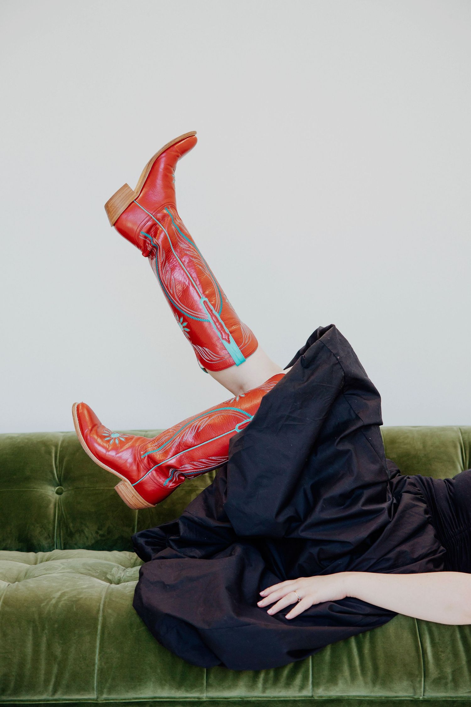 Red embroidered cowboy boots raised in air on green velvet sofa with black dress.