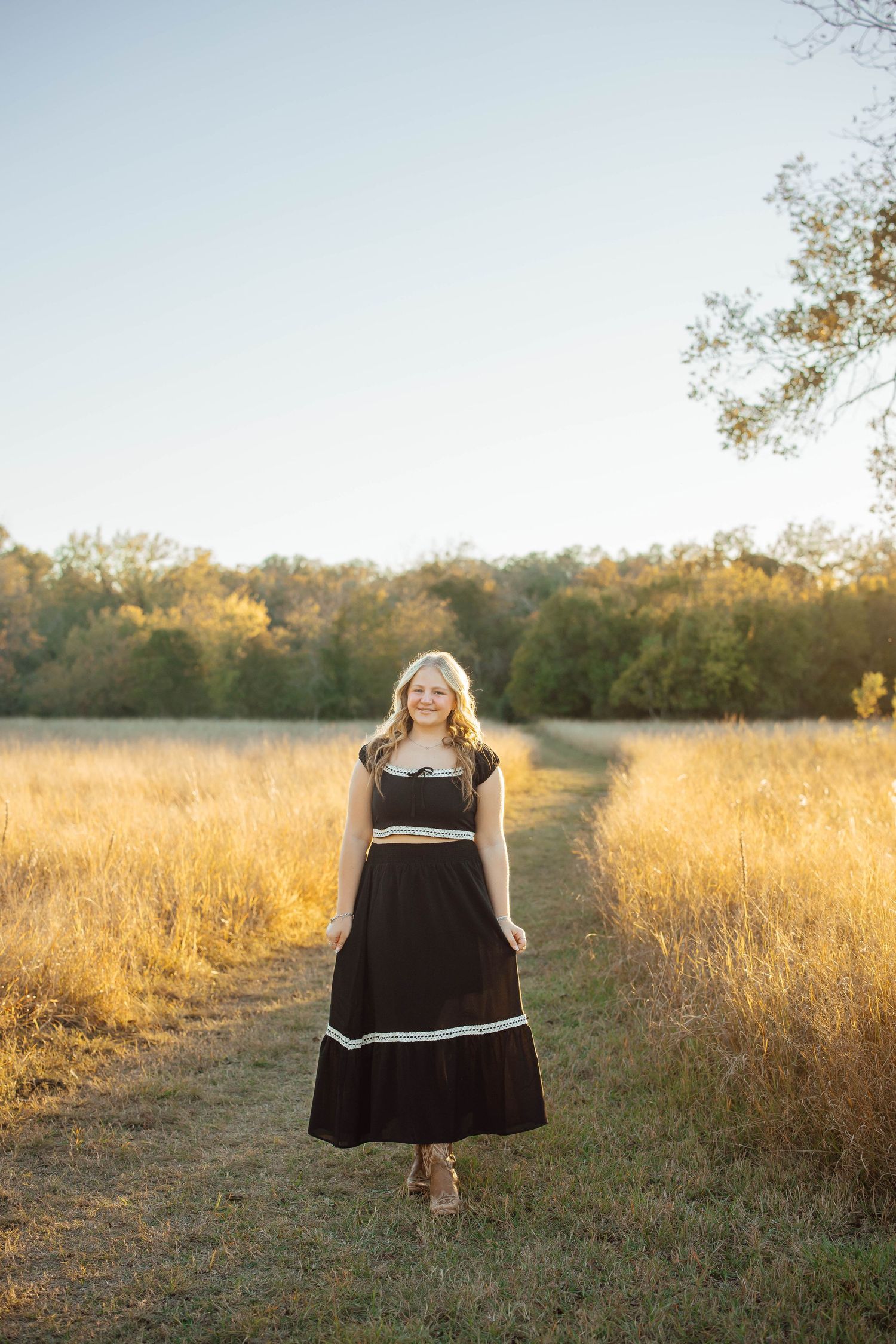 Blonde woman in black two-piece maxi outfit stands on a sunlit golden grass field path at dusk.