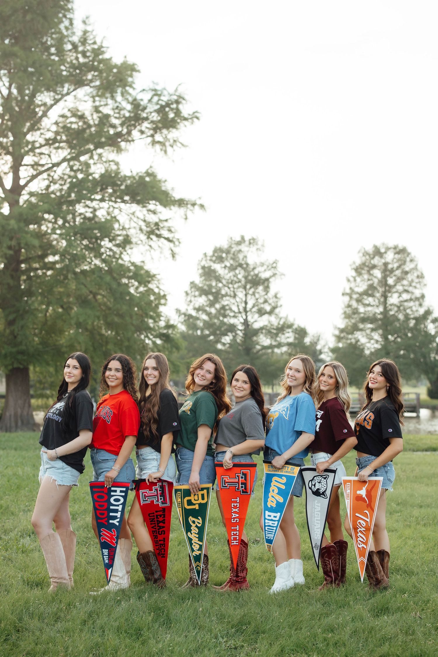 Eight young women holding college pennants pose outdoors on a green lawn.