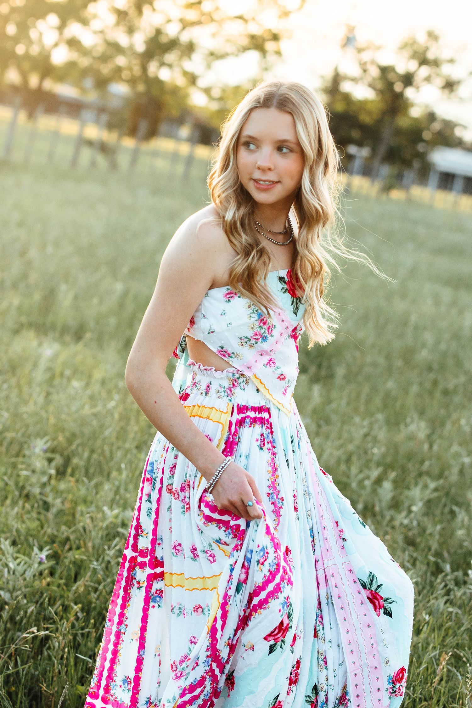 Blonde teen in colorful floral maxi dress walking through green meadow in golden sunset light.