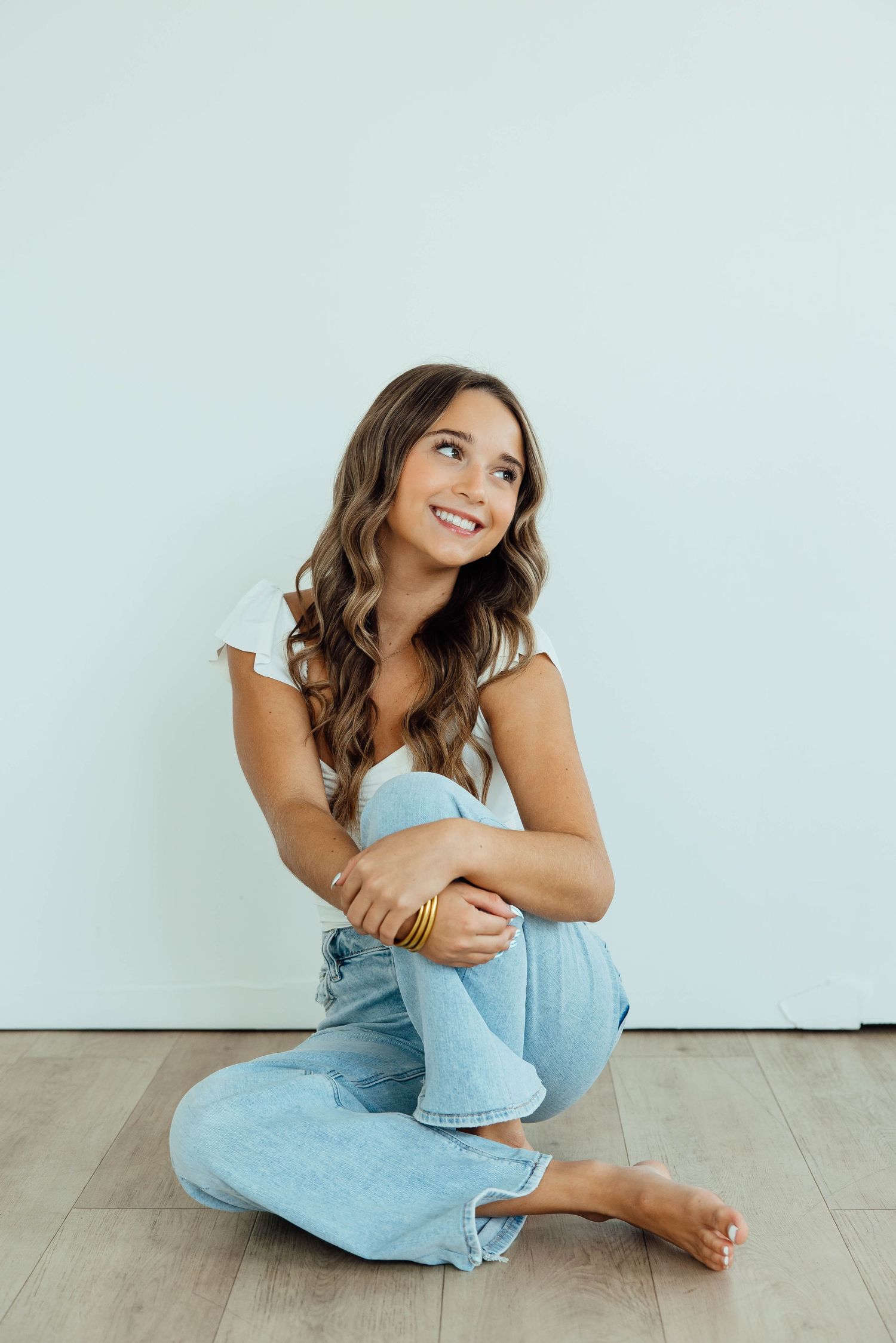 Smiling brunette woman in white top and jeans sits cross-legged on hardwood floor against white wall.