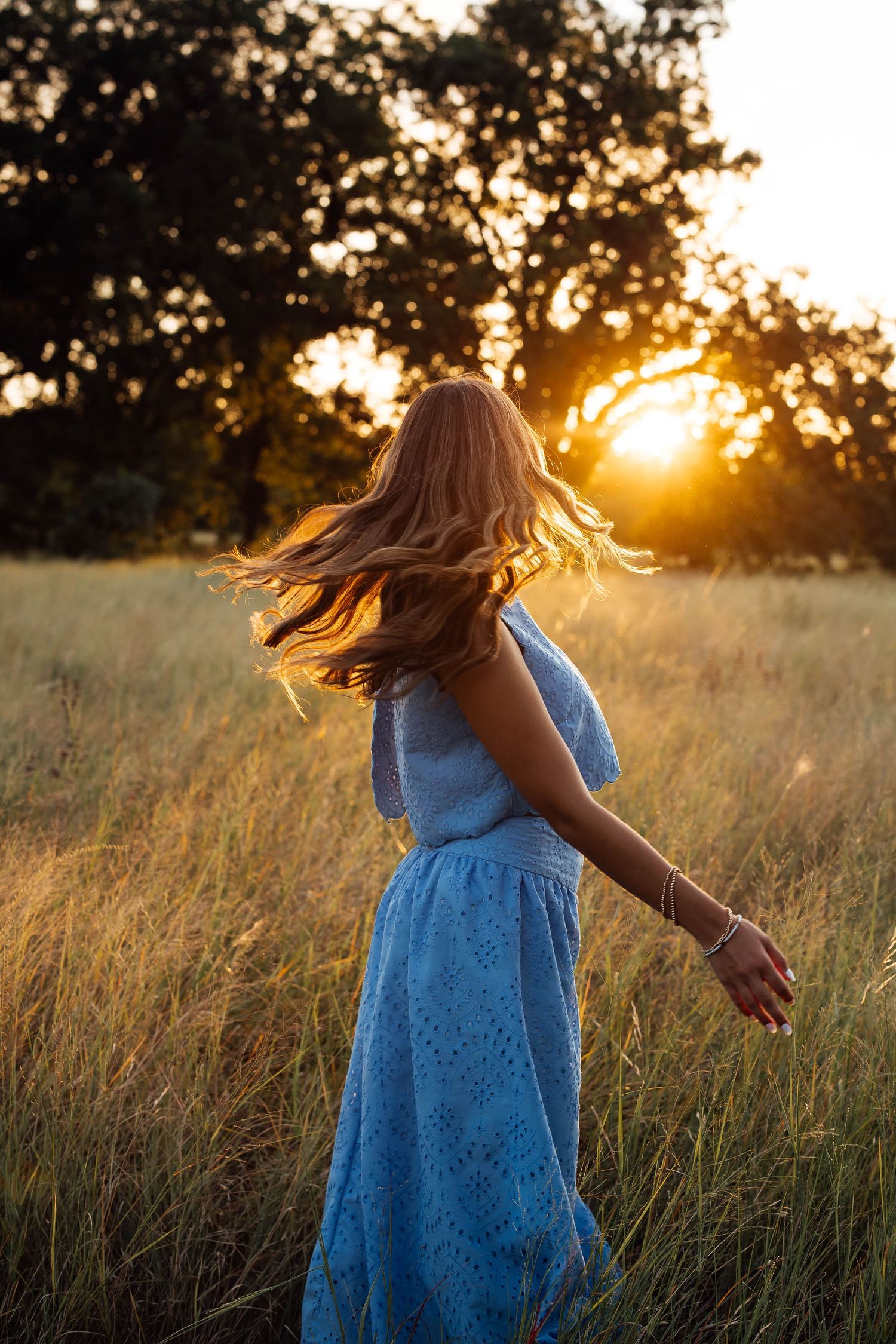 Young woman in blue eyelet dress twirling in a golden field during a stunning sunset.