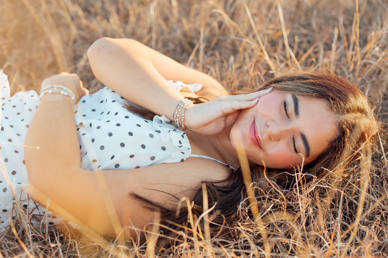 Young woman in white polka dot dress lying in golden dry grass field, smiling with eyes closed.