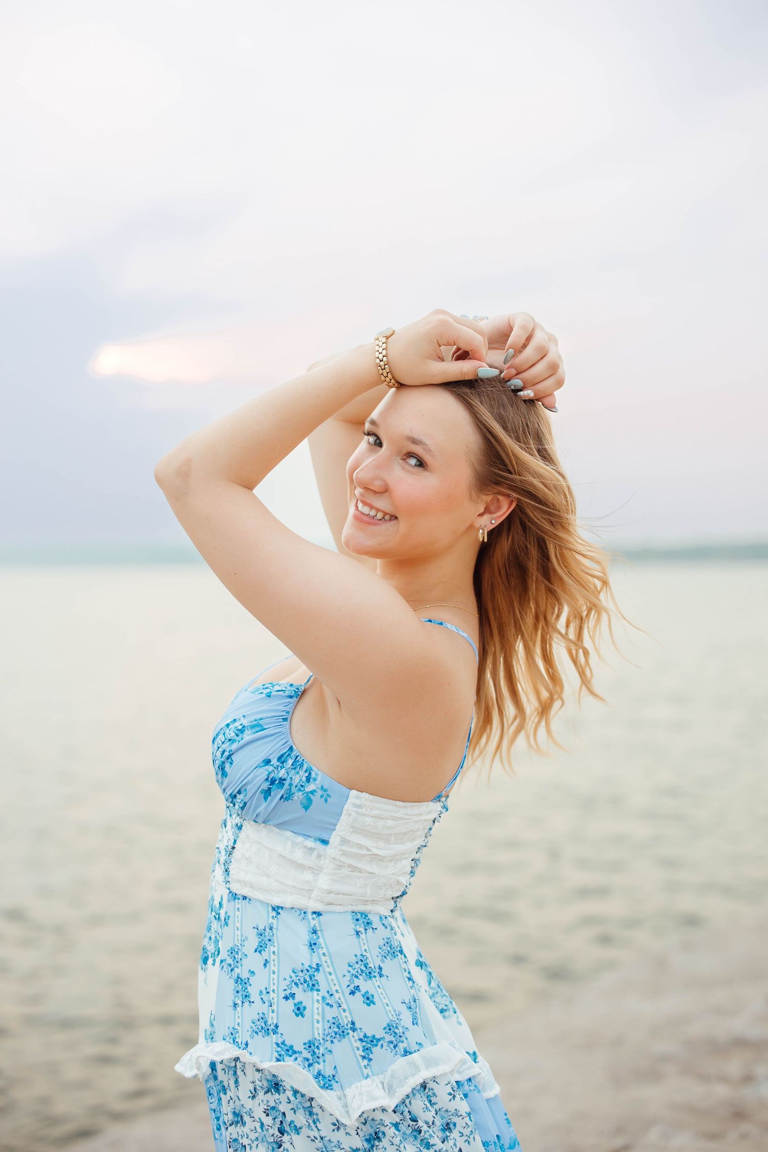 Smiling blonde woman in blue floral strapless dress poses playfully by calm lake at twilight.
