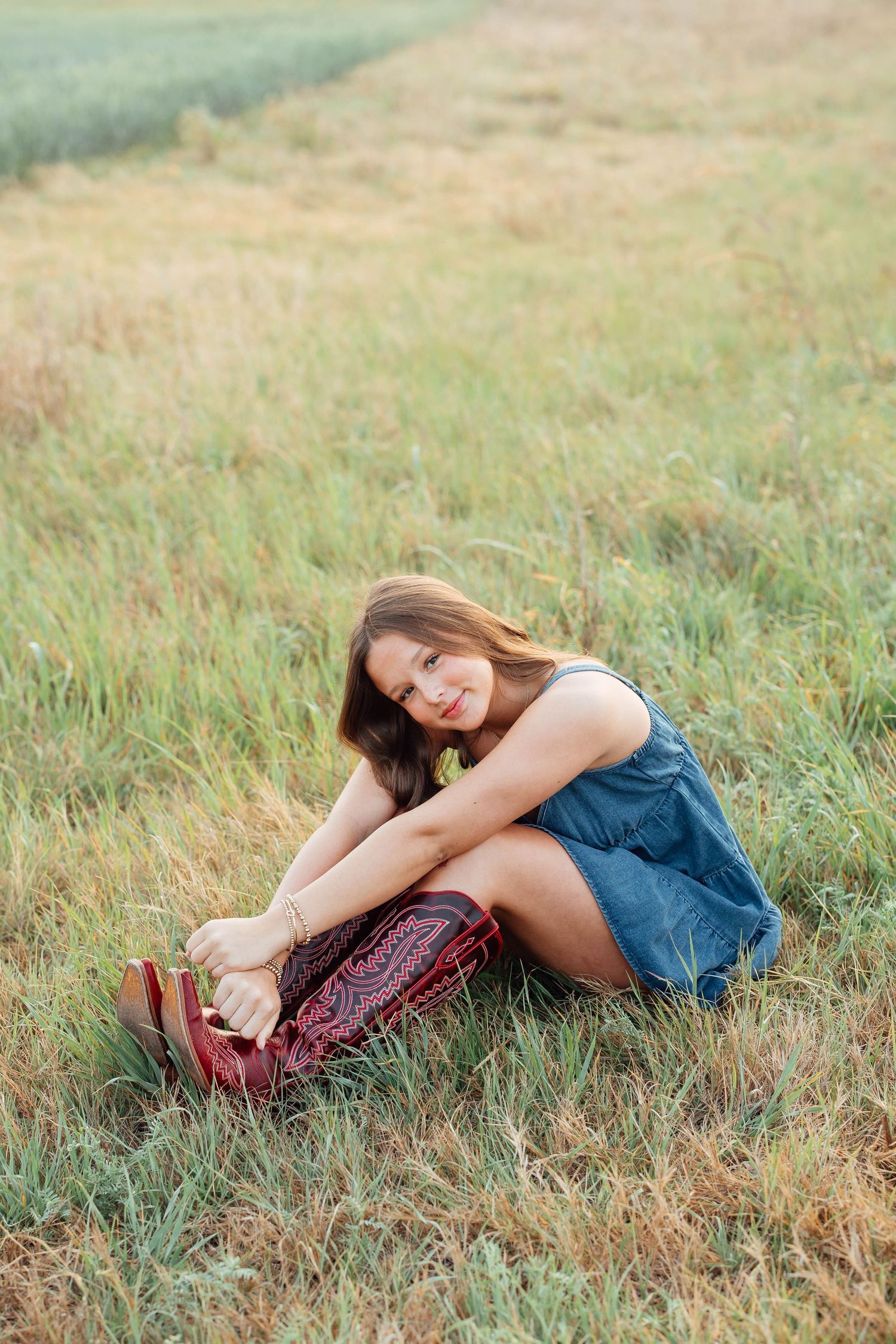 Young woman in denim dress sitting in golden grass field holding red patterned bag, smiling at camera.