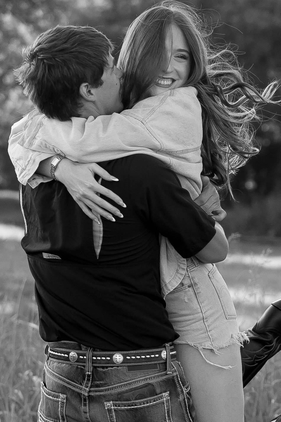 Black and white photo of laughing couple embracing in a field during golden hour outdoors.