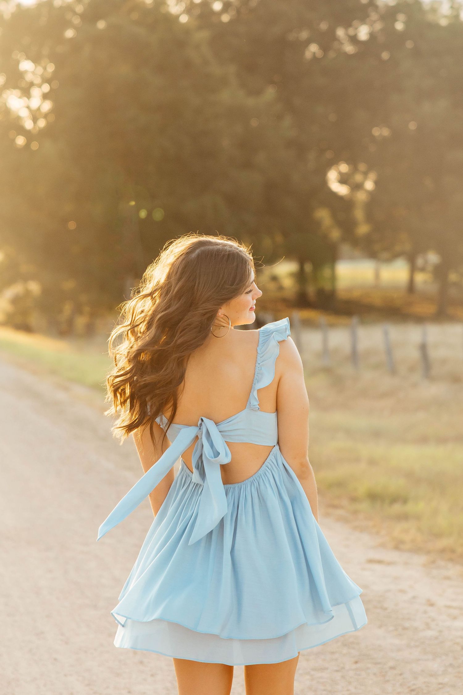 Young woman in light blue backless bow dress walks down a sunny rural dirt road at golden hour.