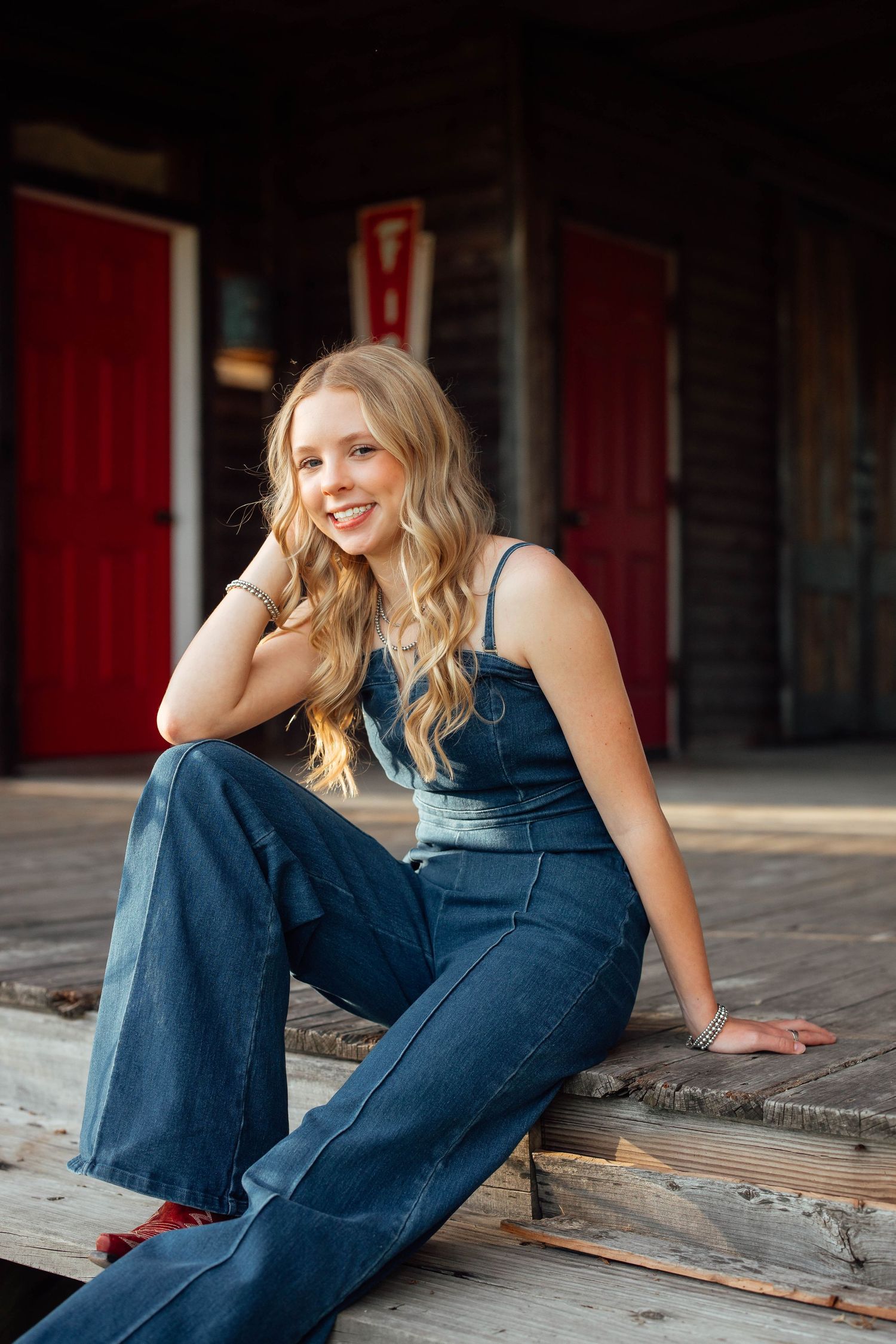 Smiling blonde teen in denim overalls sitting on rustic wooden porch steps with red barn doors.