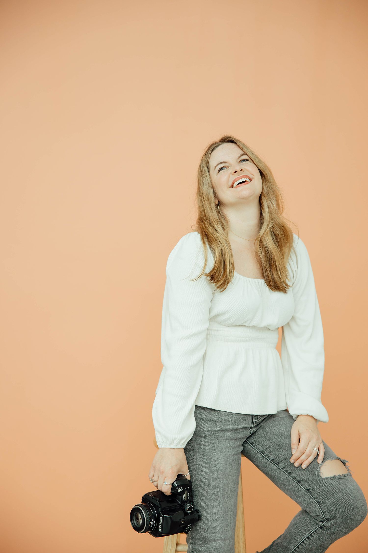 Laughing blonde woman in white blouse holding DSLR camera while seated against warm peach background.