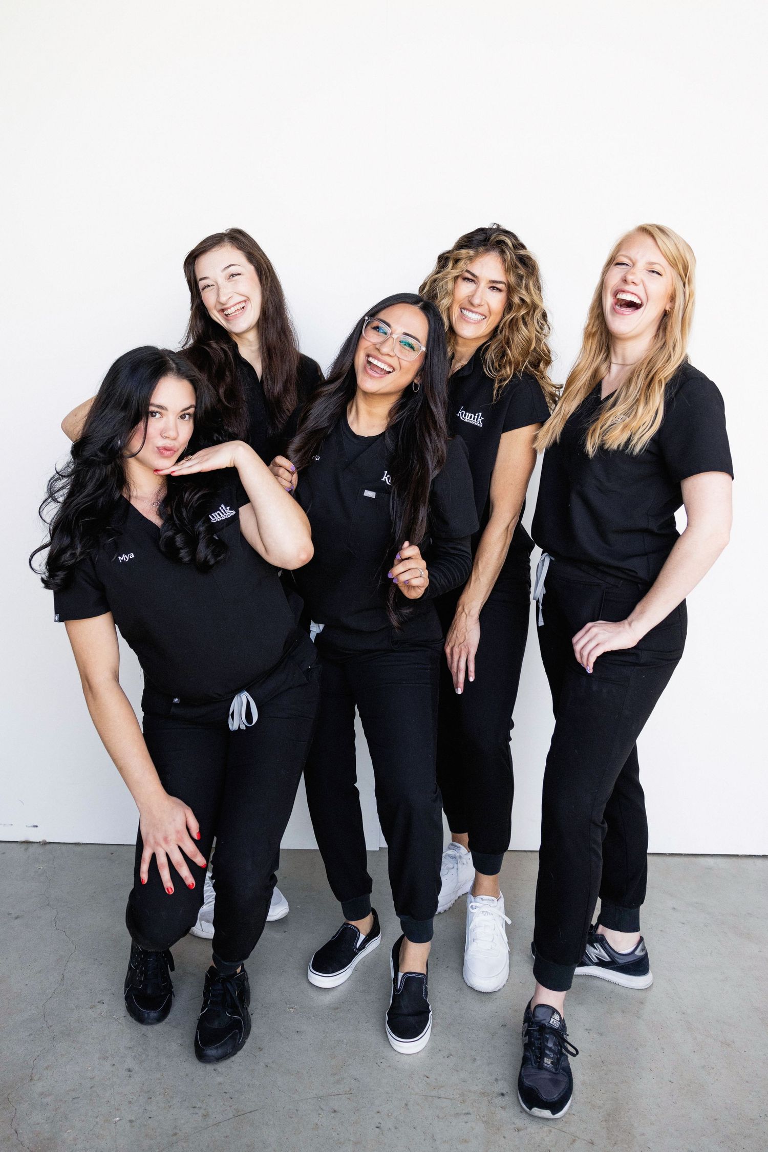 Five women in matching black scrubs uniforms smiling and laughing together against a white wall.