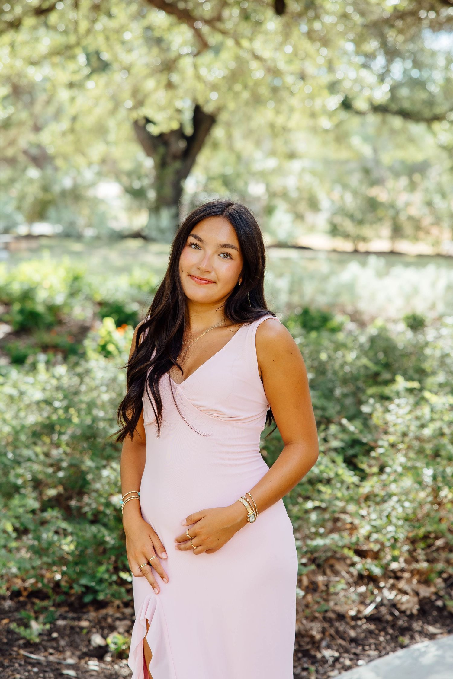 Young brunette woman in blush pink sleeveless dress poses in sunlit garden with green foliage.