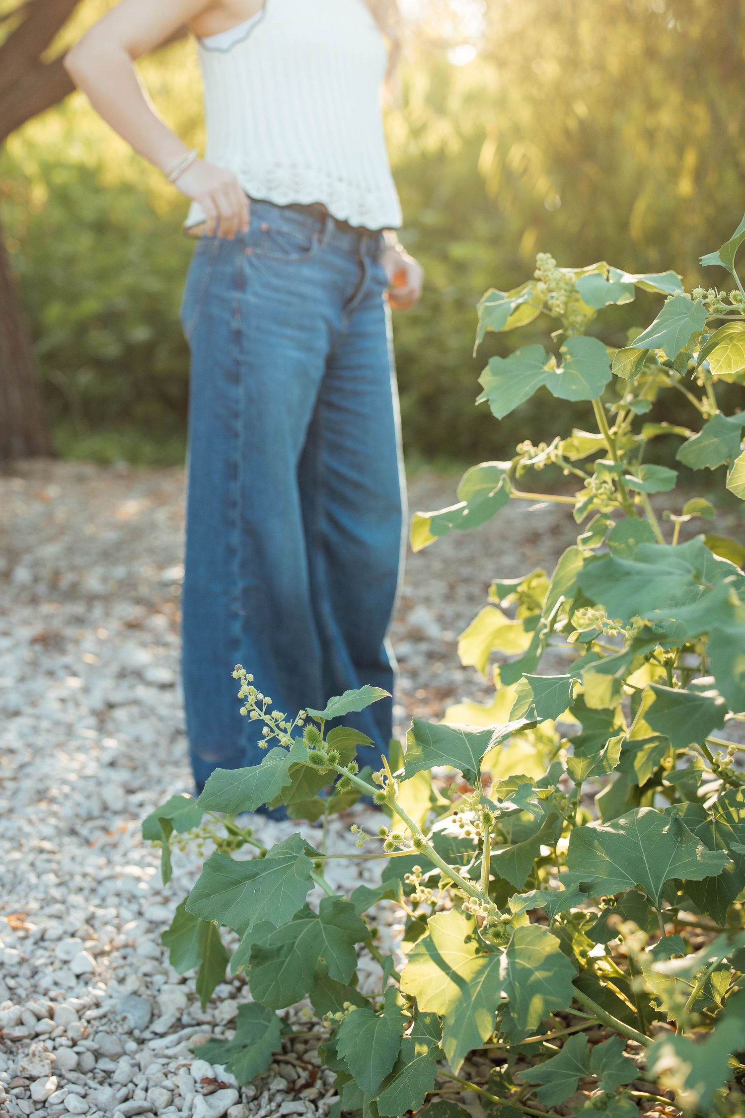 Woman in white top and wide-leg jeans standing on a gravel path surrounded by green foliage.
