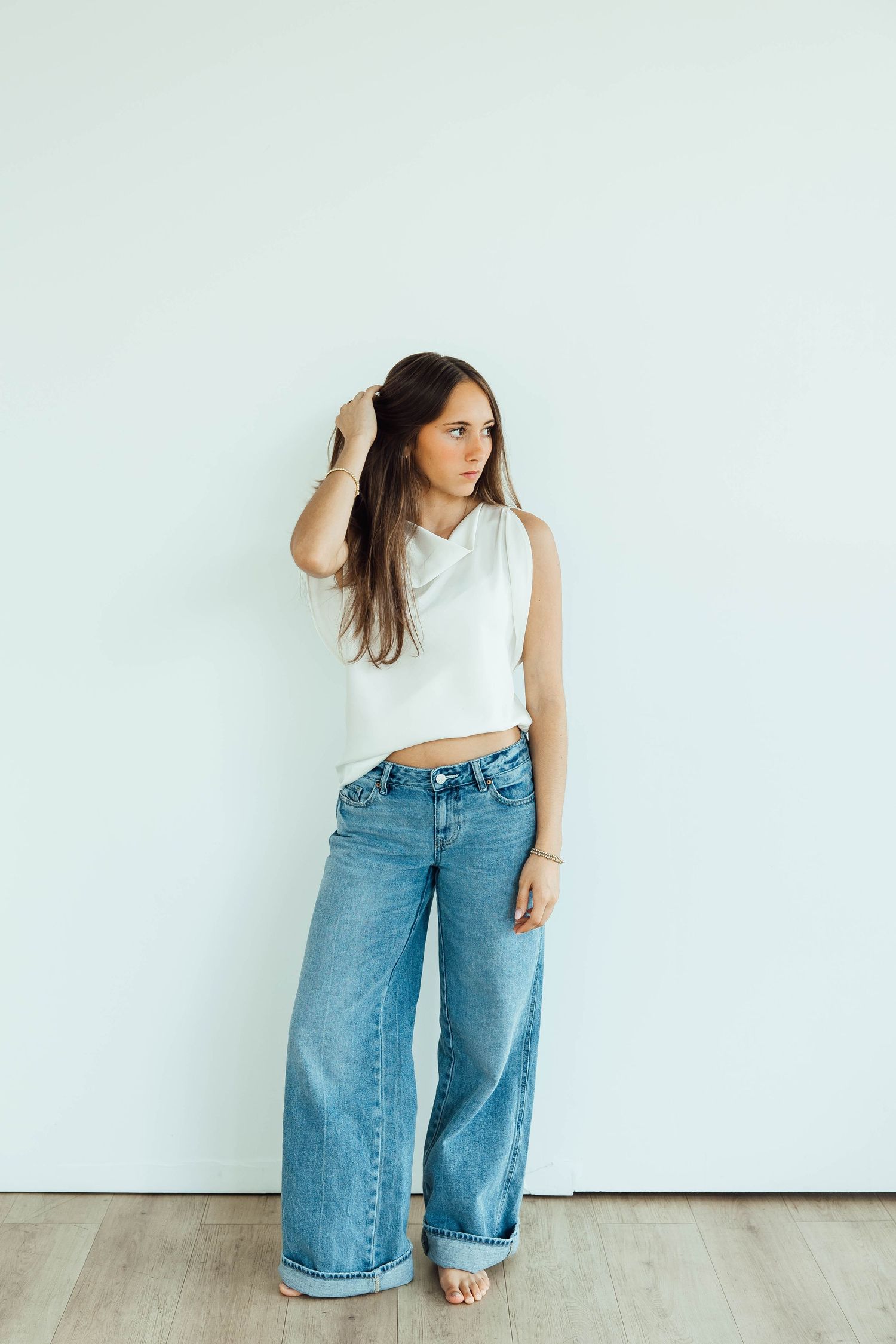 Brunette woman in white sleeveless top and wide-leg jeans posing against a light gray wall.