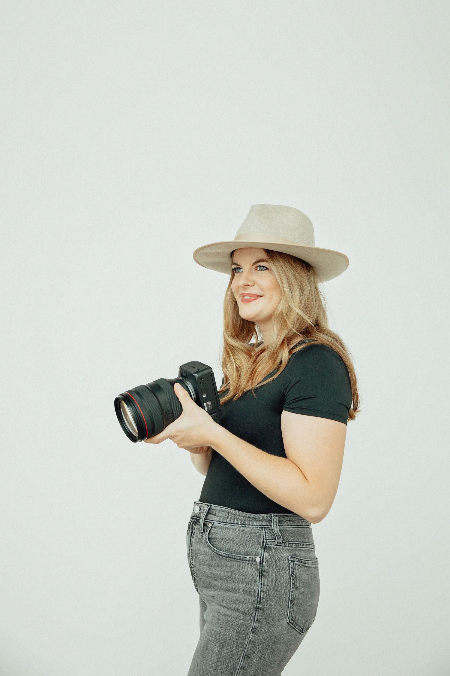 Smiling blonde woman in black top and hat holding professional DSLR camera against light grey background.