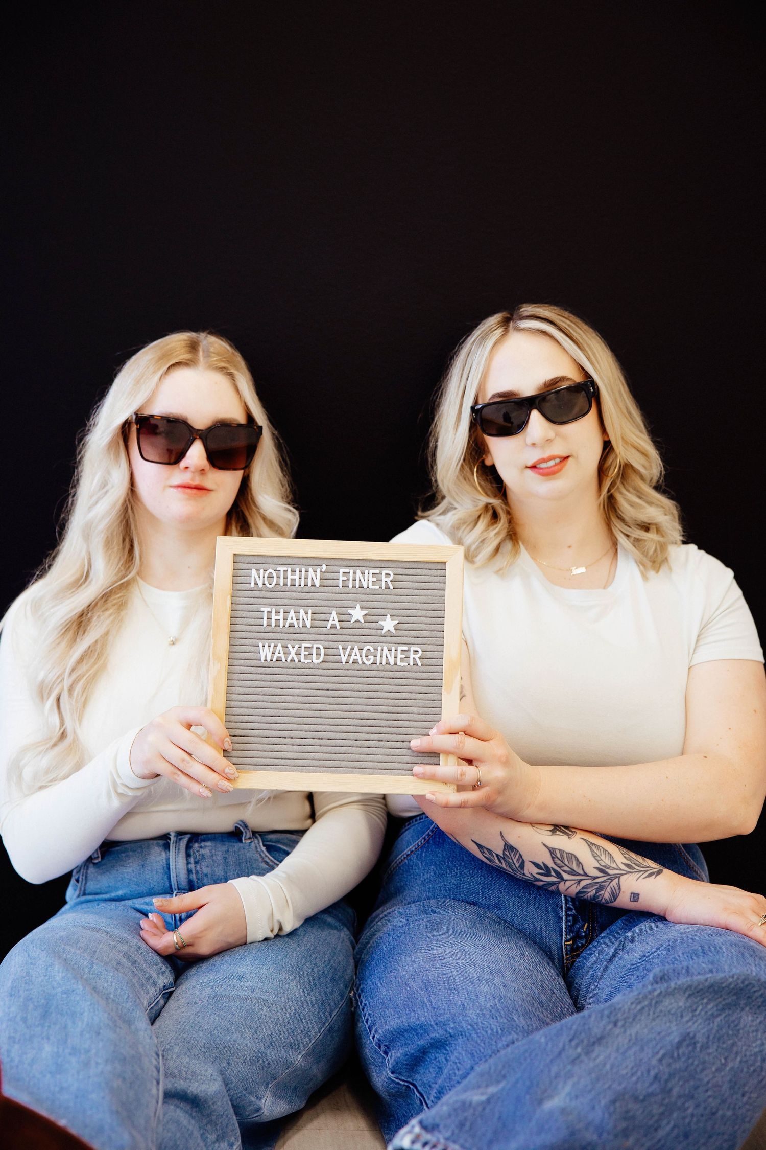 Two blonde women in sunglasses holding letterboard sign on black background.