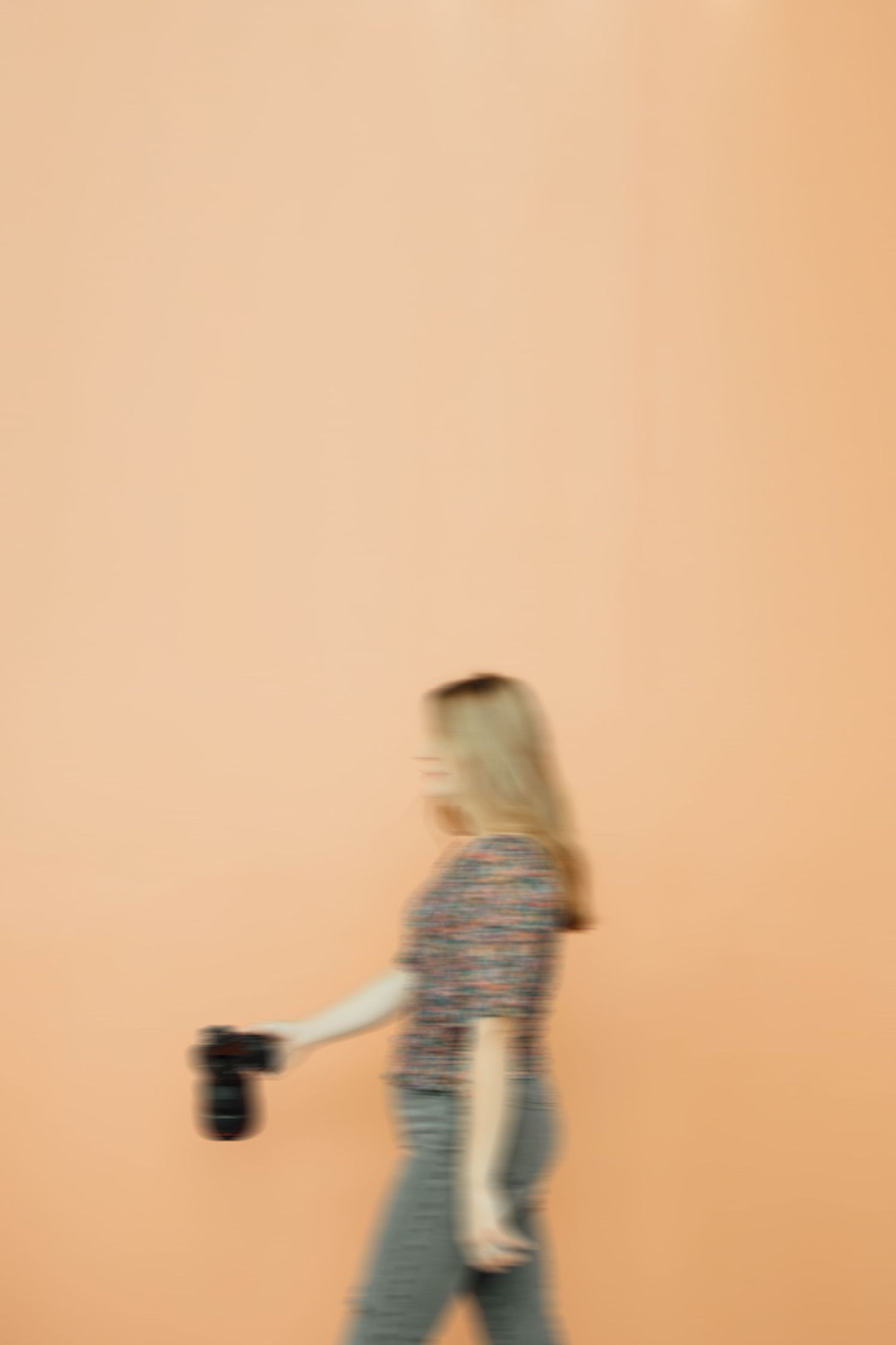 Motion blur artistic photo of blonde woman walking with camera against warm orange background.