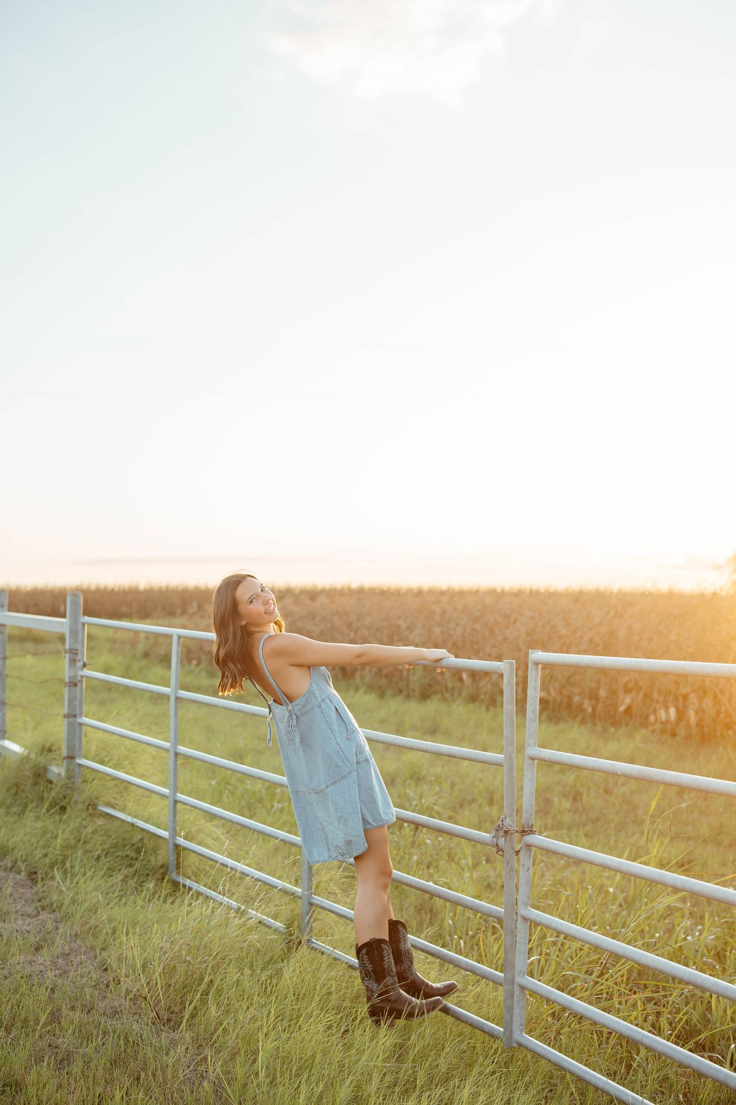 Young woman in blue dress and cowboy boots leaning on a white farm fence at golden hour sunset.