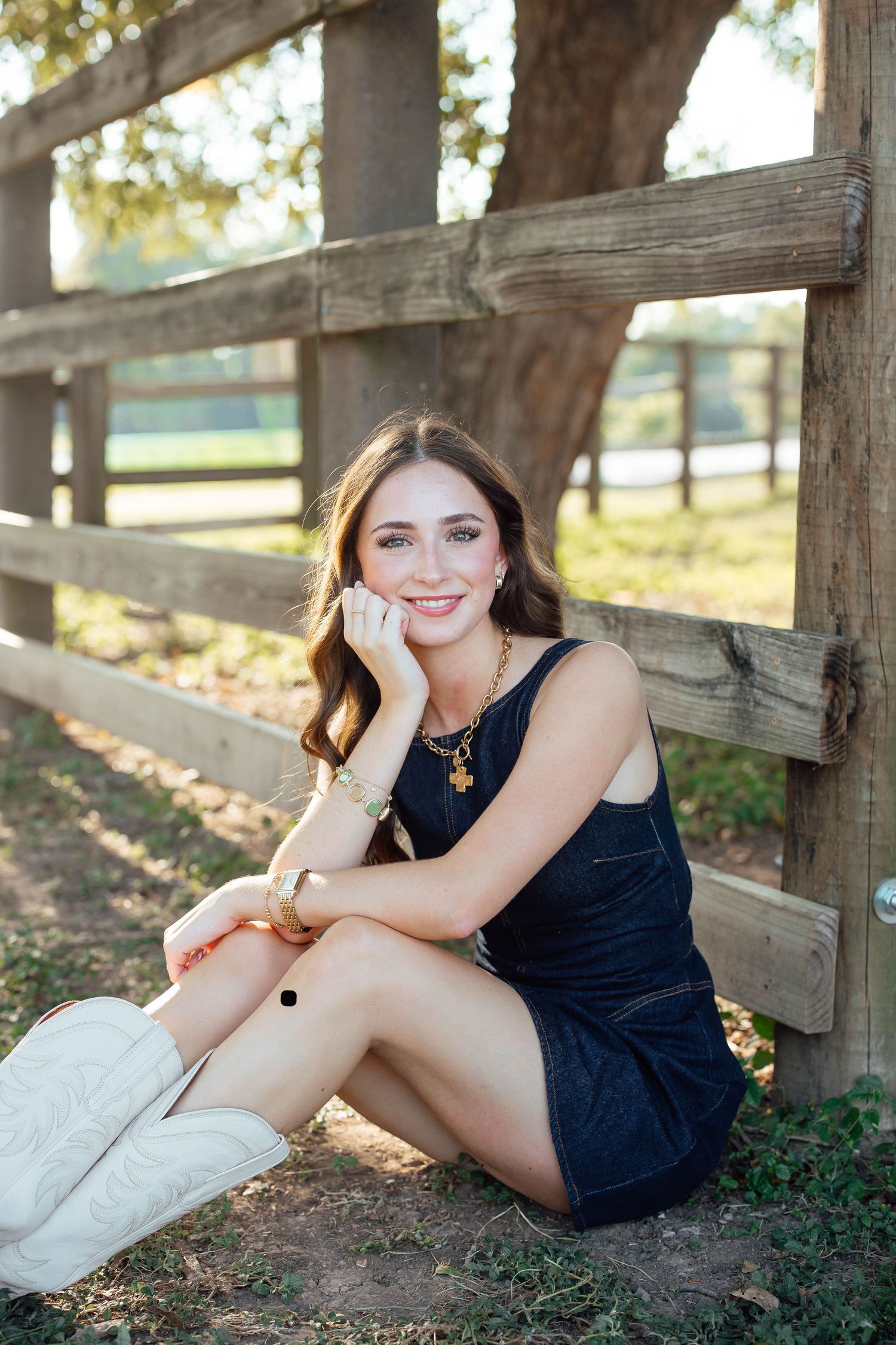 A smiling young woman in a black dress and white cowboy boots sits against a wooden fence outdoors.
