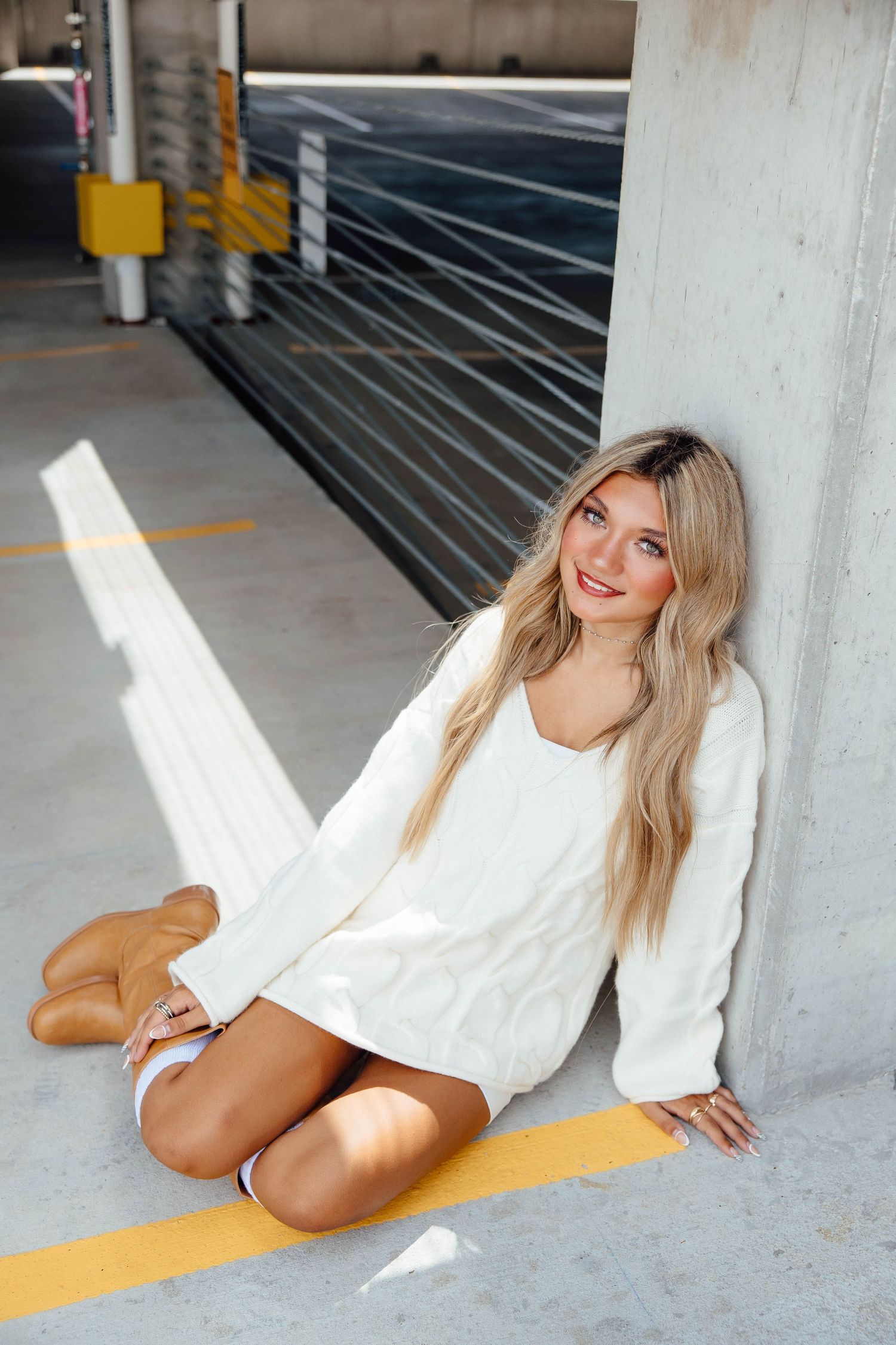 Smiling blonde woman in white oversized sweater sits on parking garage floor against concrete pillar.