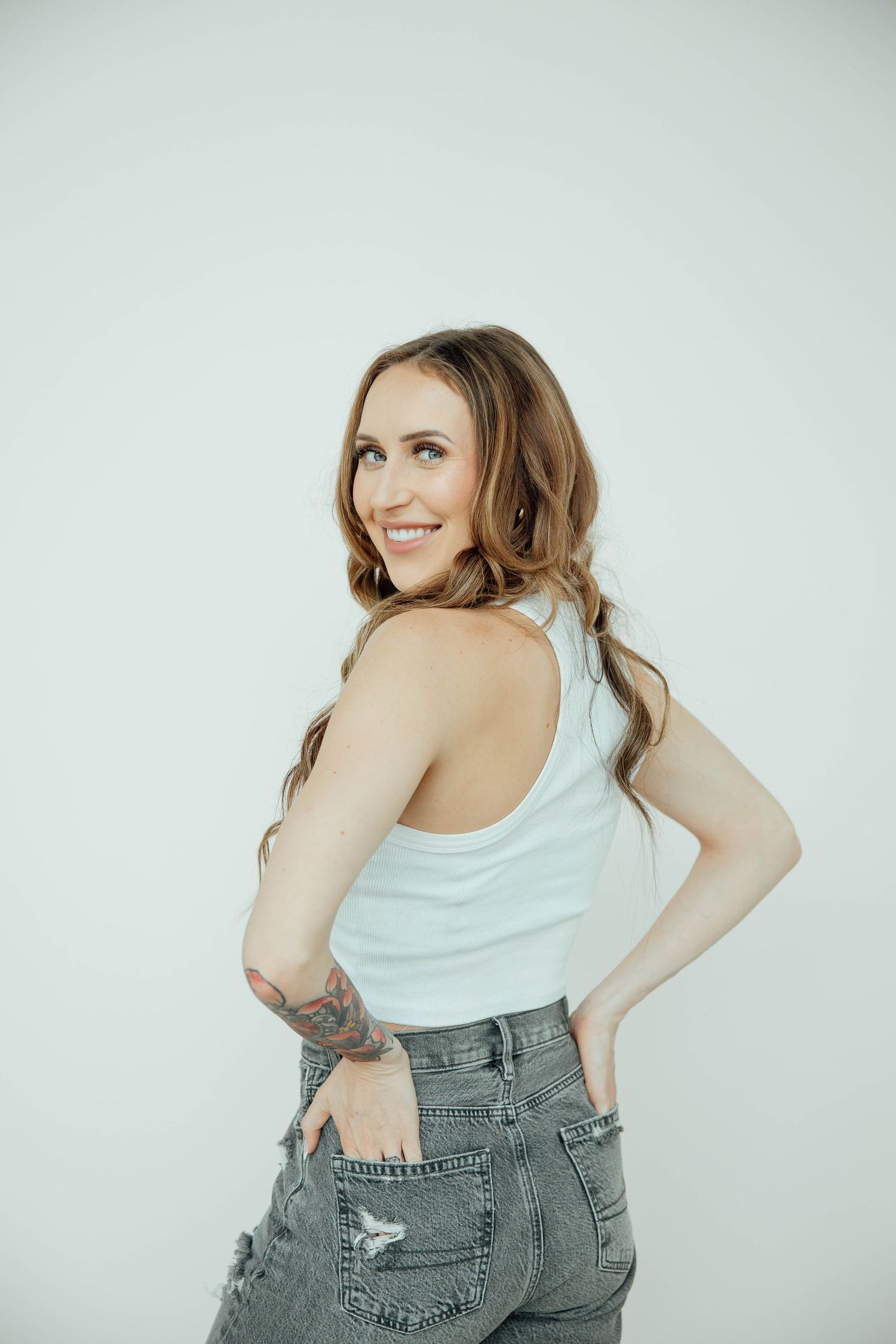 Smiling brunette woman with tattoo in white tank top and grey distressed jeans posing against white background.