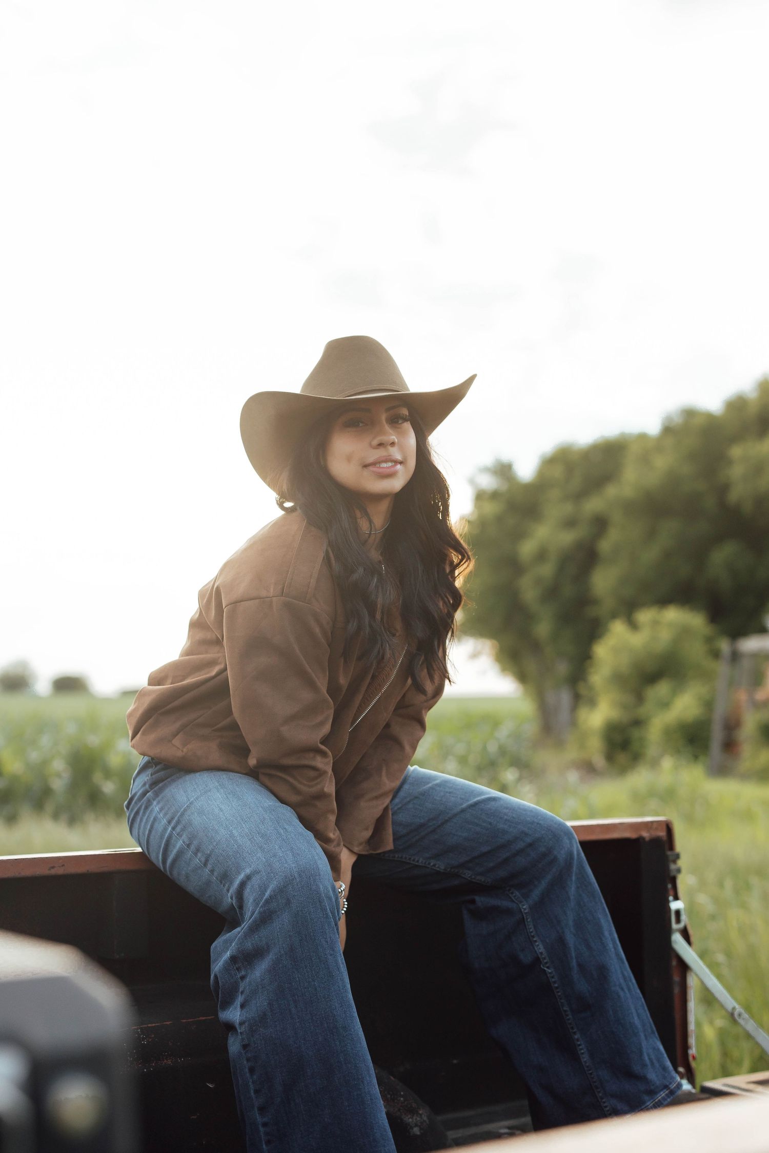 A woman in a brown jacket and jeans sits on a truck tailgate wearing a dark cowboy hat outdoors.