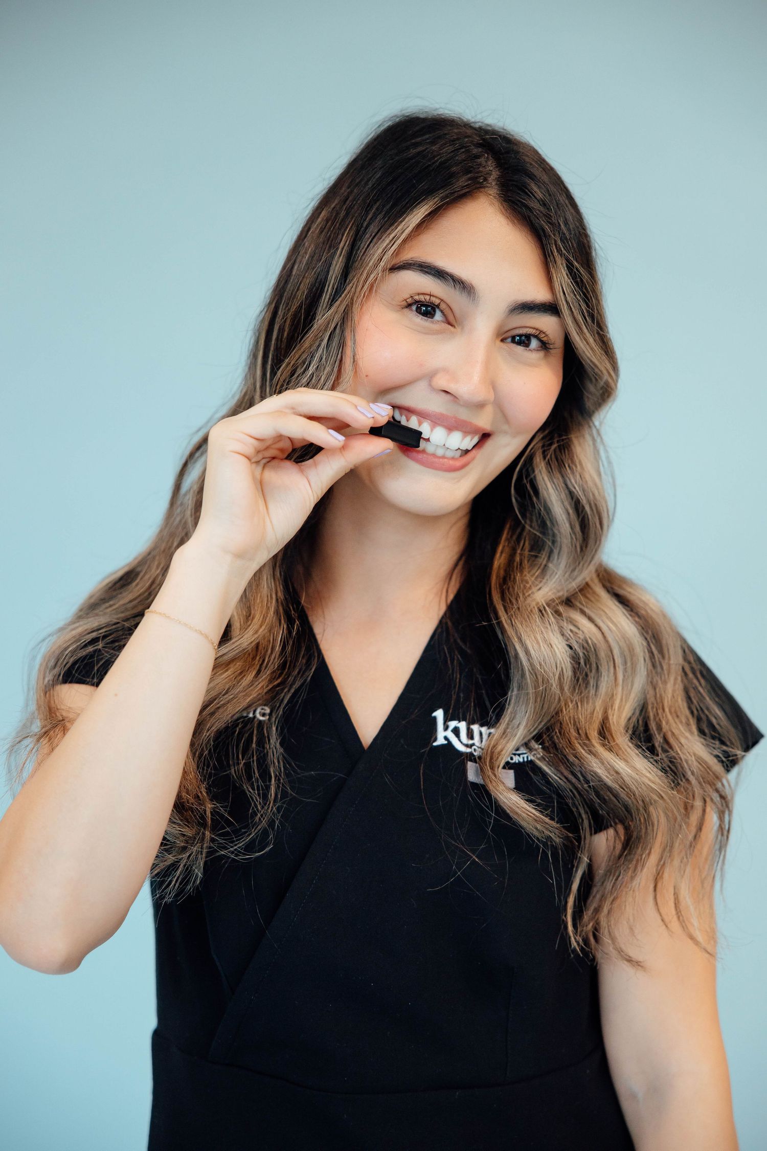 Brunette woman in black scrubs smiling while holding a clear dental aligner on light blue background.