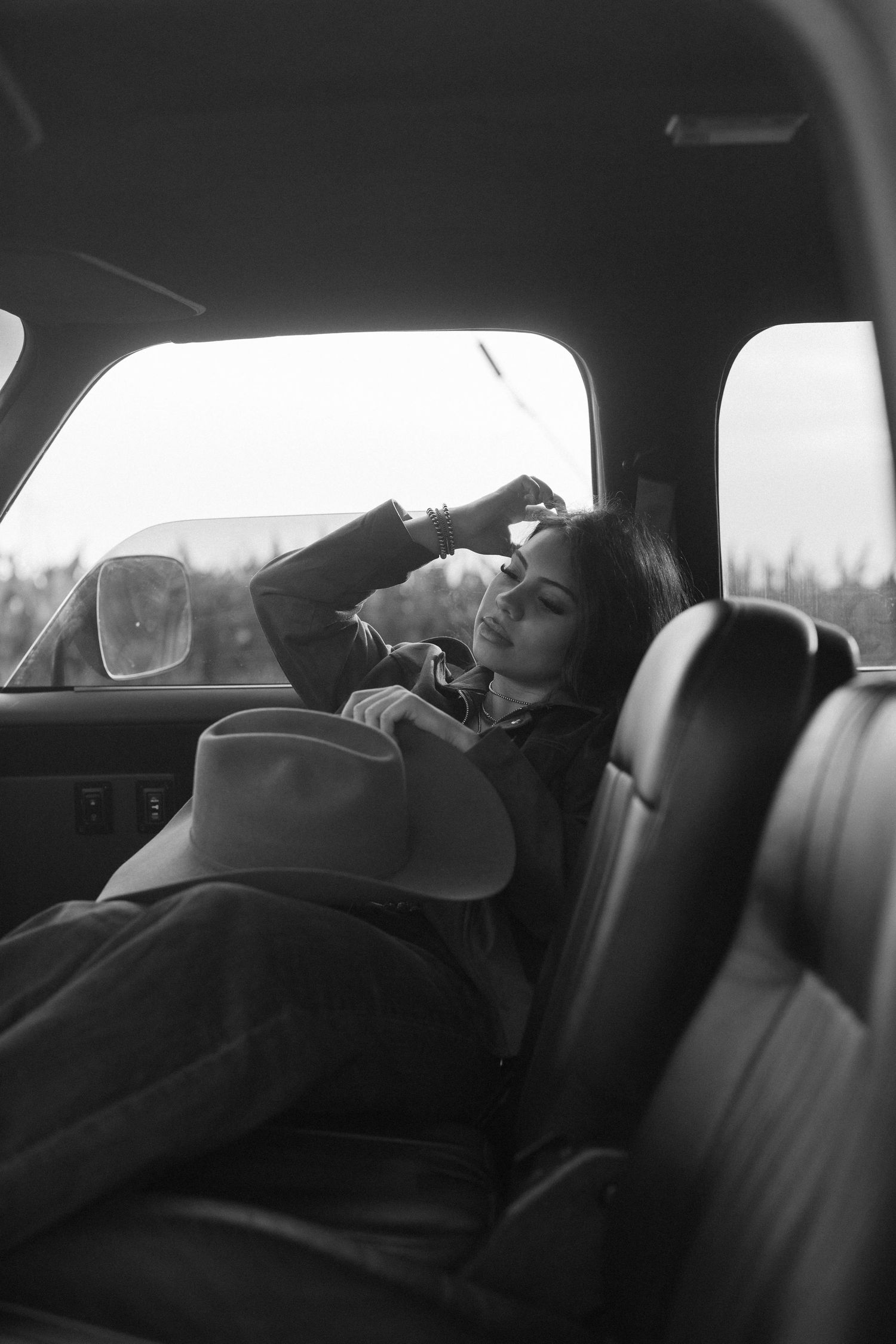 Black and white photo of a woman in a cowboy hat reclining in the back seat of a vintage vehicle.