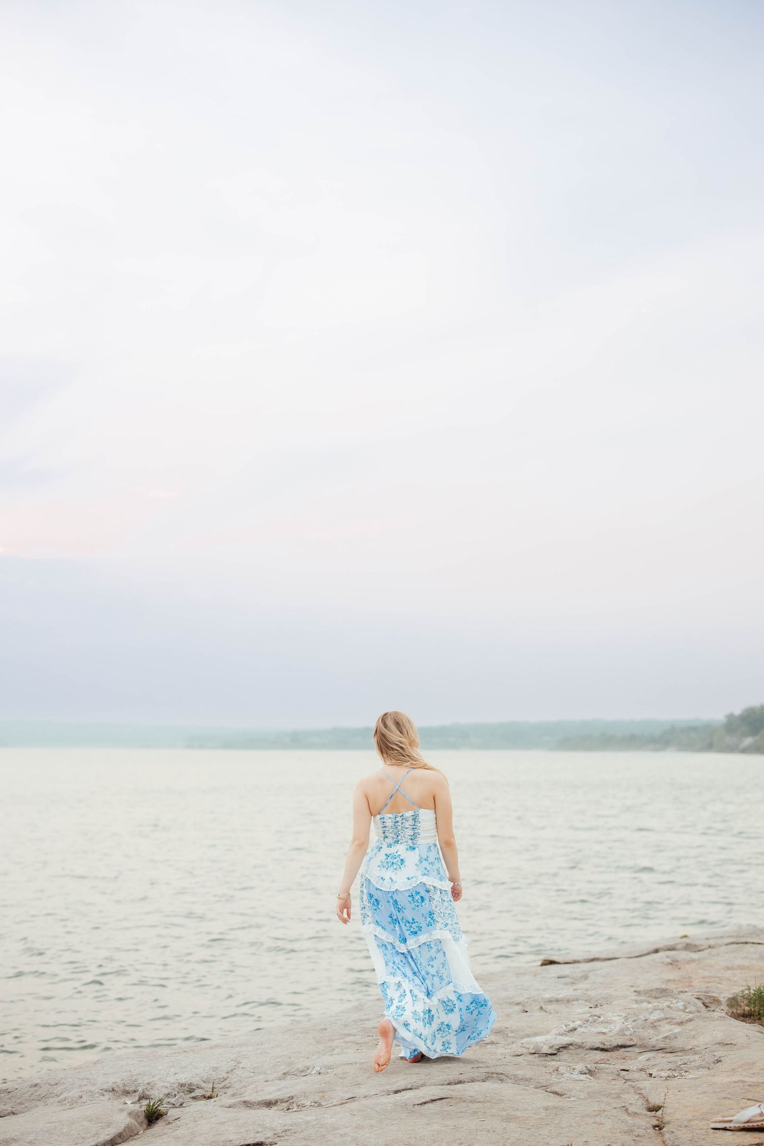 Blonde woman in blue floral maxi dress stands barefoot on rocky lakeside shore under cloudy sky.