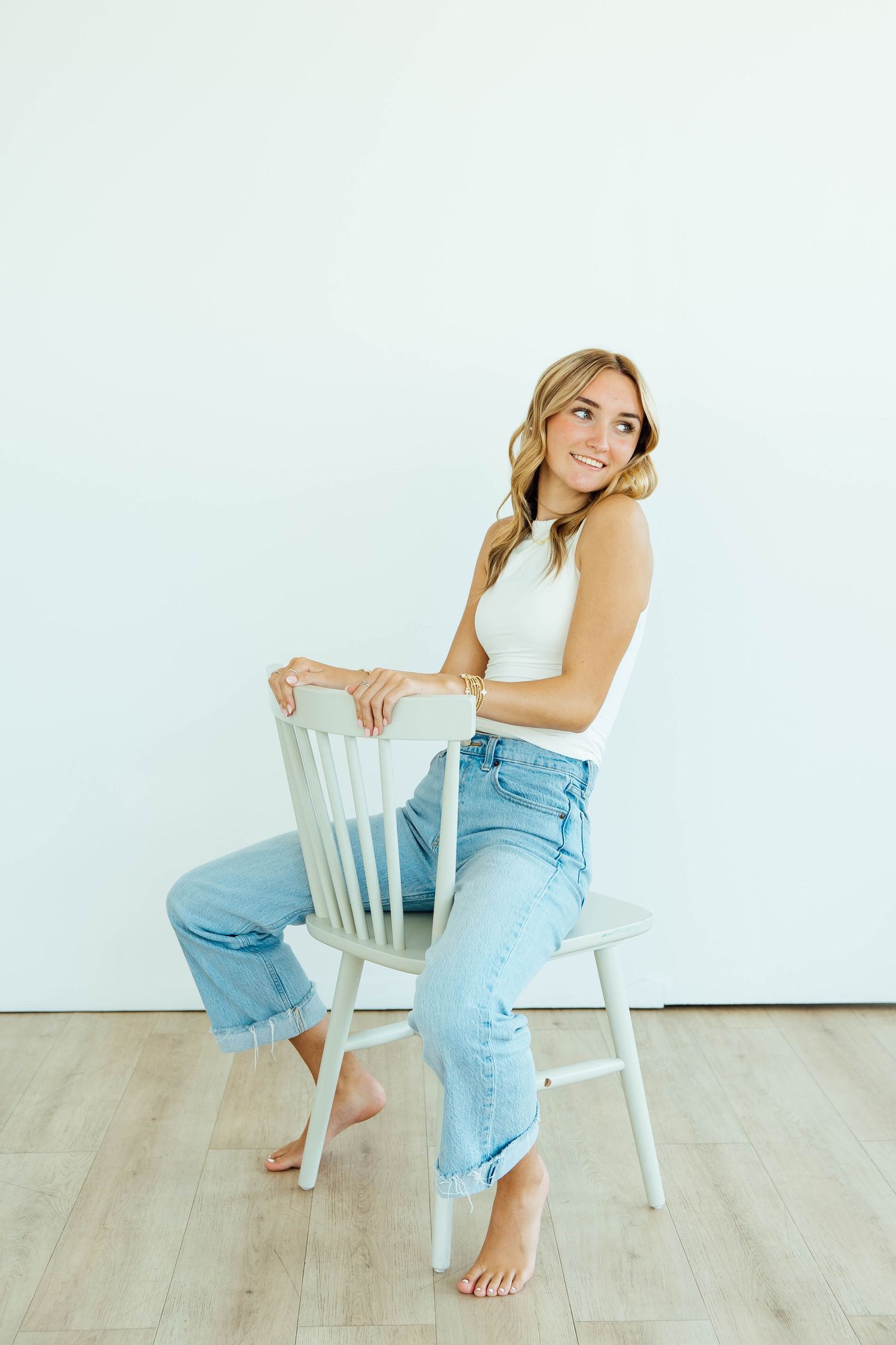 Smiling blonde woman in white sleeveless top and jeans sits sideways on white wooden chair.