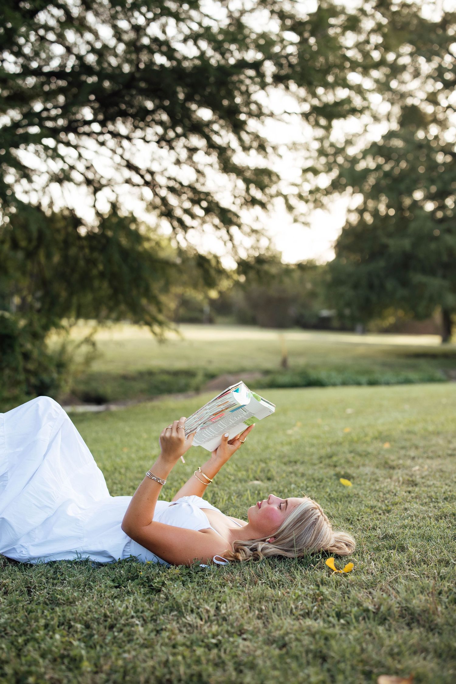 Young blonde woman in white dress lying on green grass reading a colorful book outdoors.